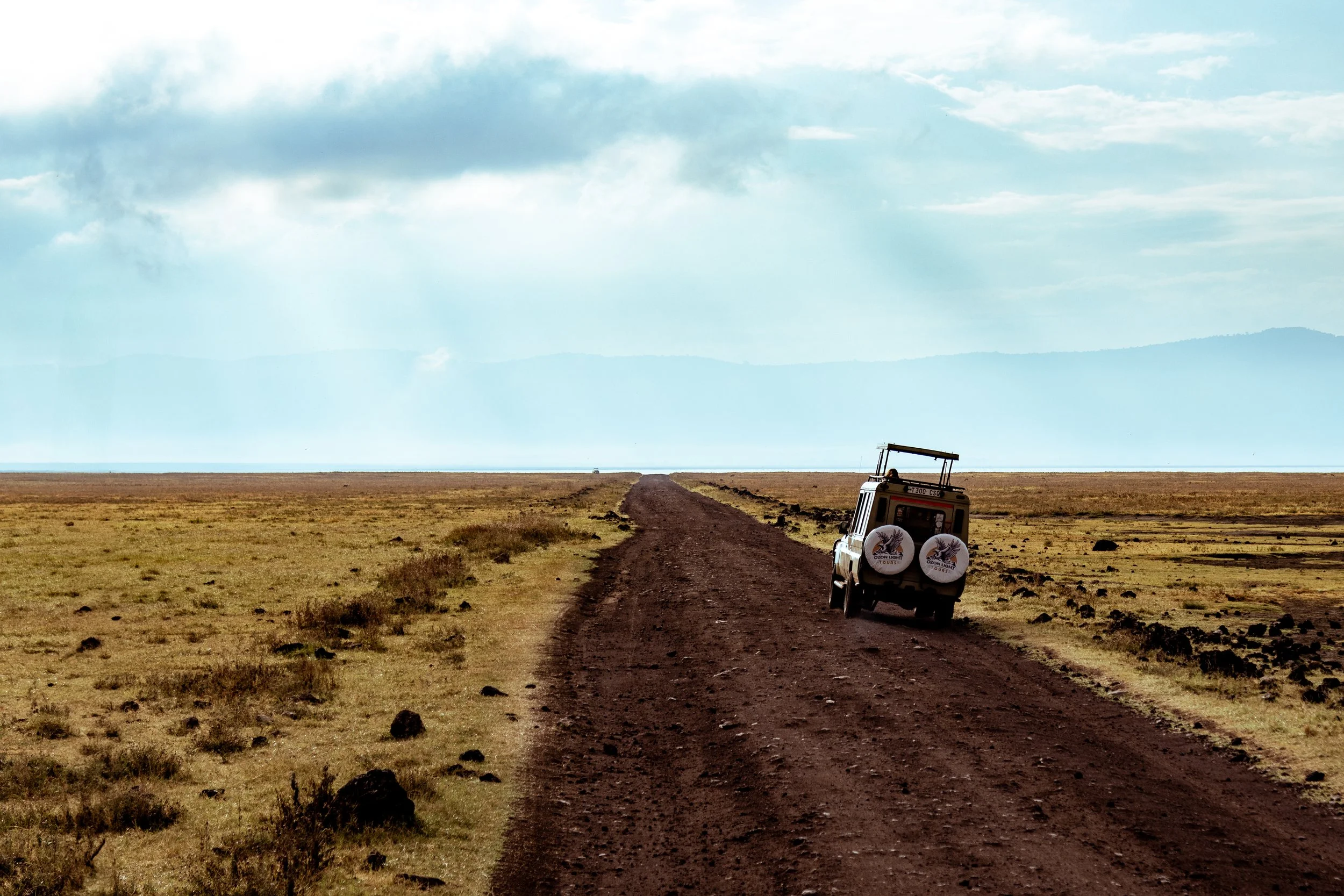 A dirt road stretching across a grassy plain with a small safari vehicle traveling on it. The landscape features wide open space and mountains in the distance, with a cloudy sky overhead.