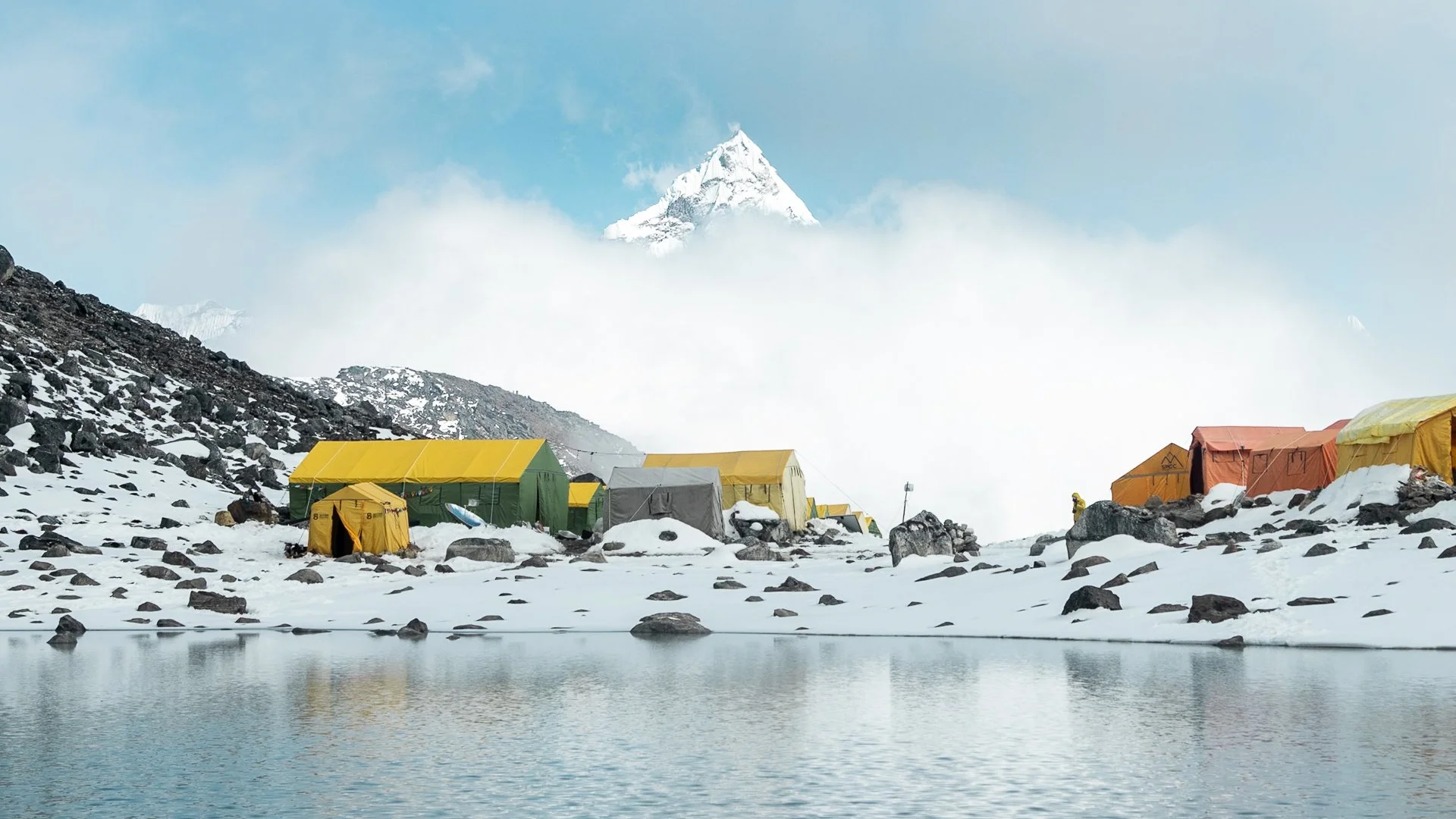 Snow-covered mountain landscape with a cluster of colorful tents on a rocky terrain near a body of water, with snow-capped peaks and a partly cloudy sky in the background.