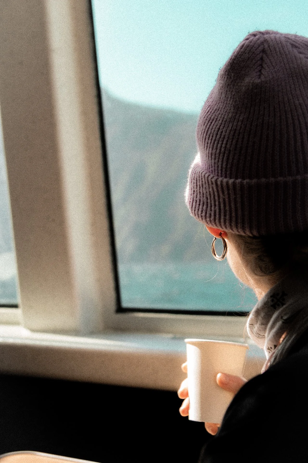 A person with a purple beanie and hoop earrings looking out a airplane window while holding a white coffee cup.