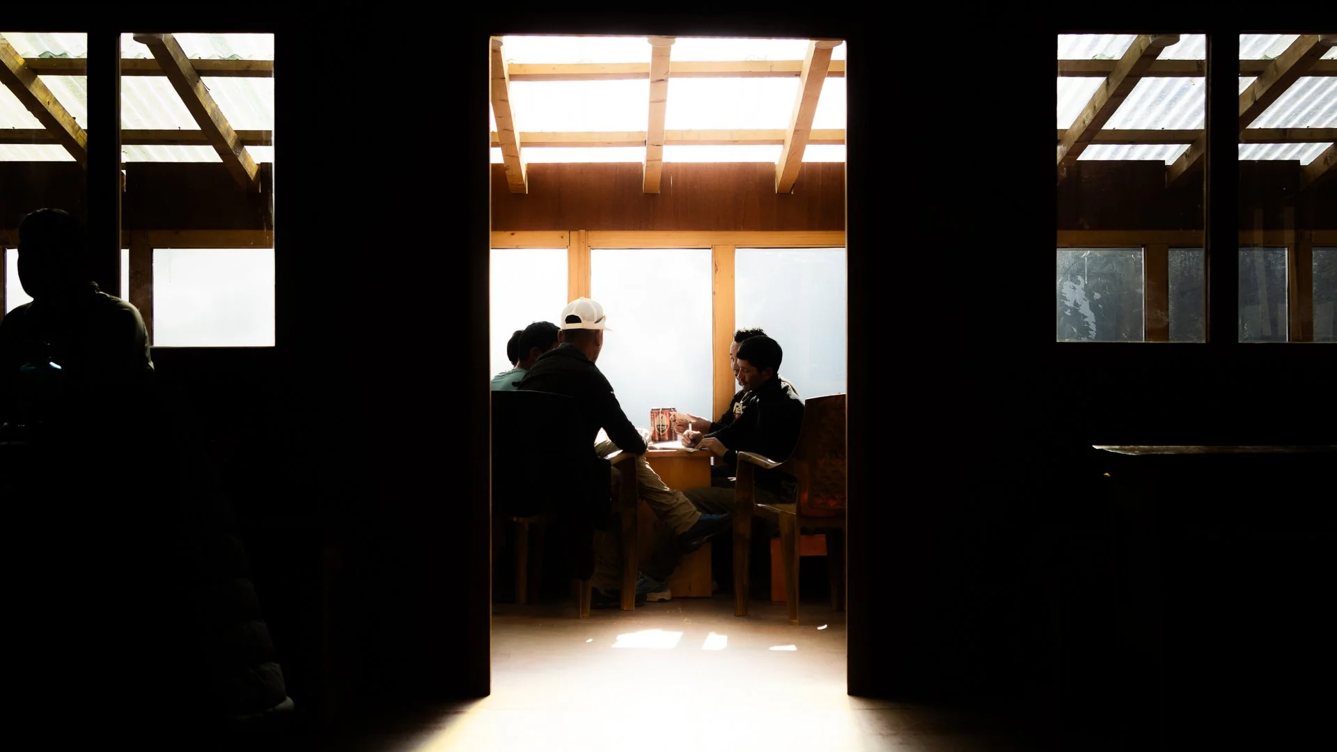 People sitting at a table in a sunlit room with large windows, viewed through a doorway silhouetted in darkness.