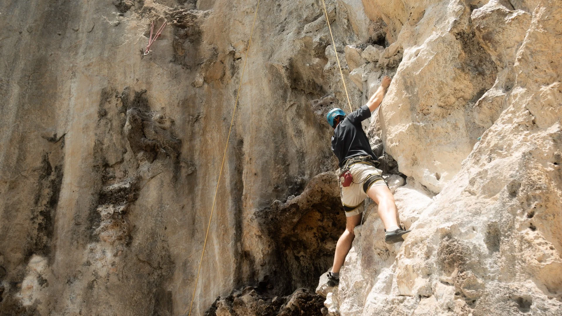 A person wearing a helmet and climbing gear ascending a steep rock face in a natural outdoor setting.