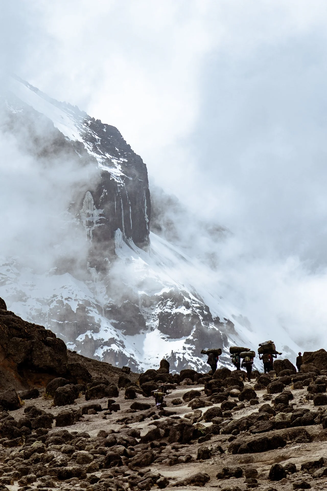 Group of hikers with backpacks walking on rocky terrain in a snow-covered mountain landscape with mist, snow, and clouds.