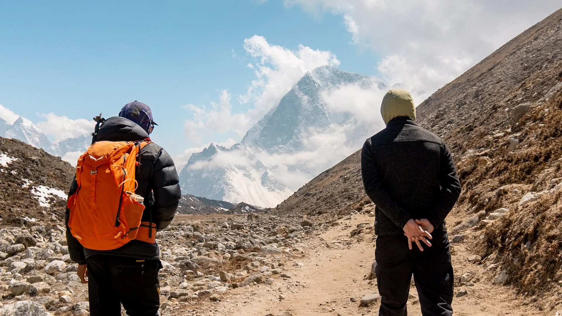 Two climbers or hikers standing on a rocky mountain trail, with snow-capped peaks and cloudy sky in the background.