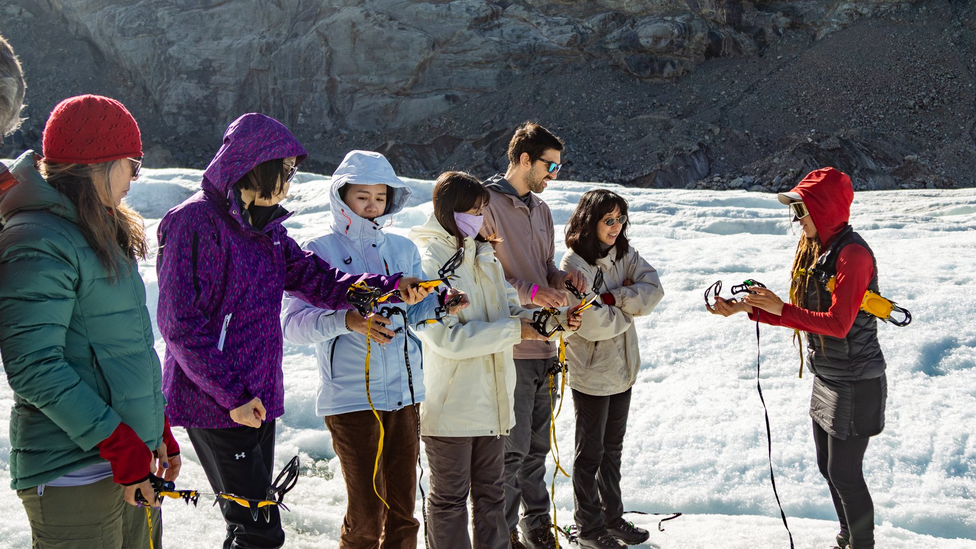 Group of people in winter clothing standing on snow, preparing for a snowshoe activity, with rocky terrain in the background.