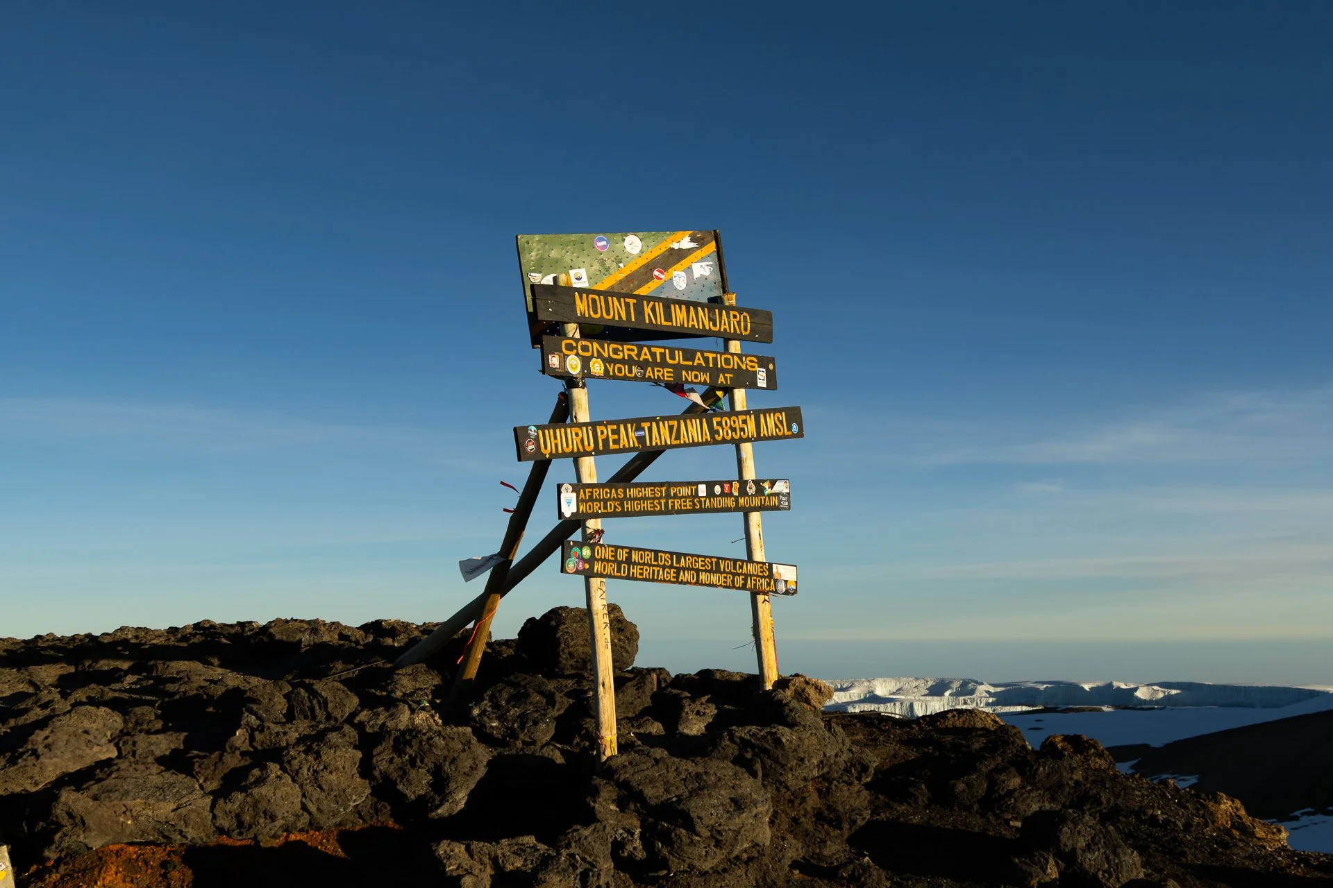 Sign at Mount Kilimanjaro indicating the summit at 5,895 meters above sea level, with additional facts about the mountain on a clear day with volcanic rocks and distant snow-capped peaks.