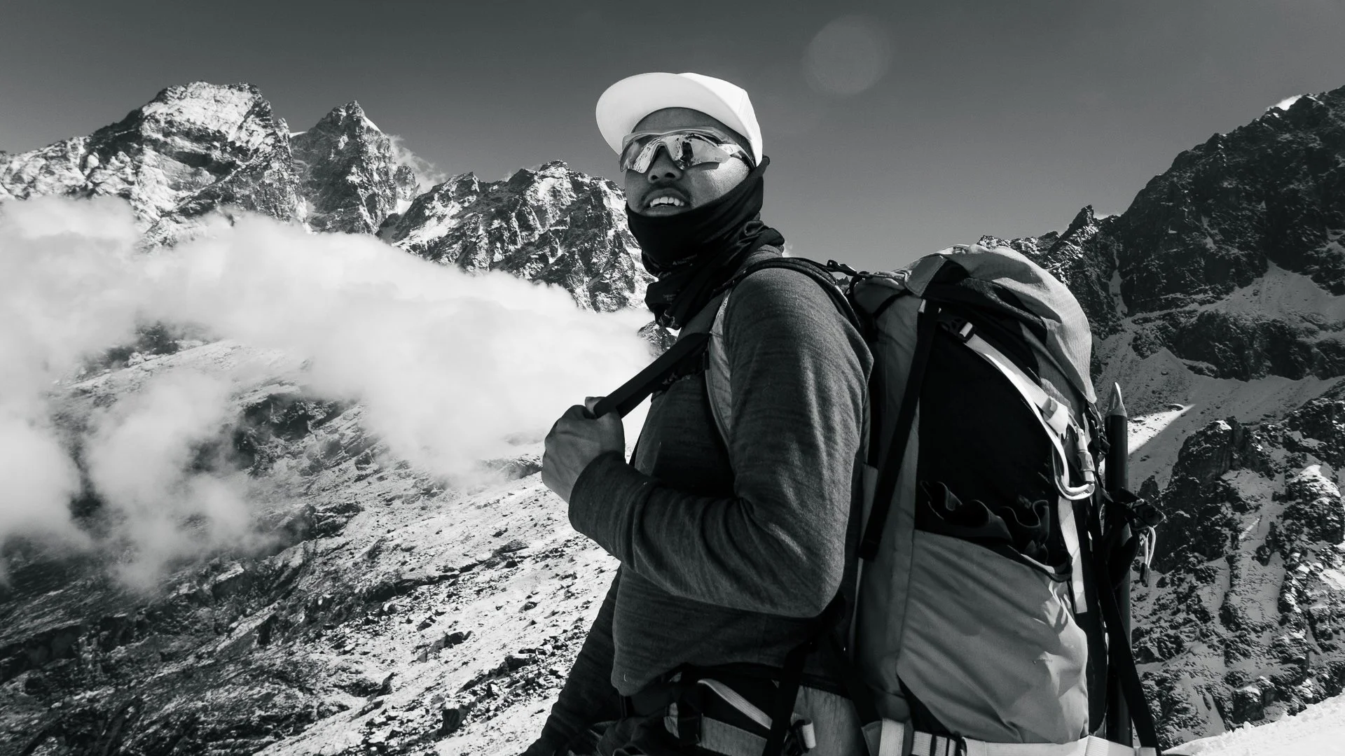 A person in outdoor winter gear with sunglasses, a cap, and a backpack, standing in a snowy mountainous landscape with rugged, snow-covered peaks in the background.