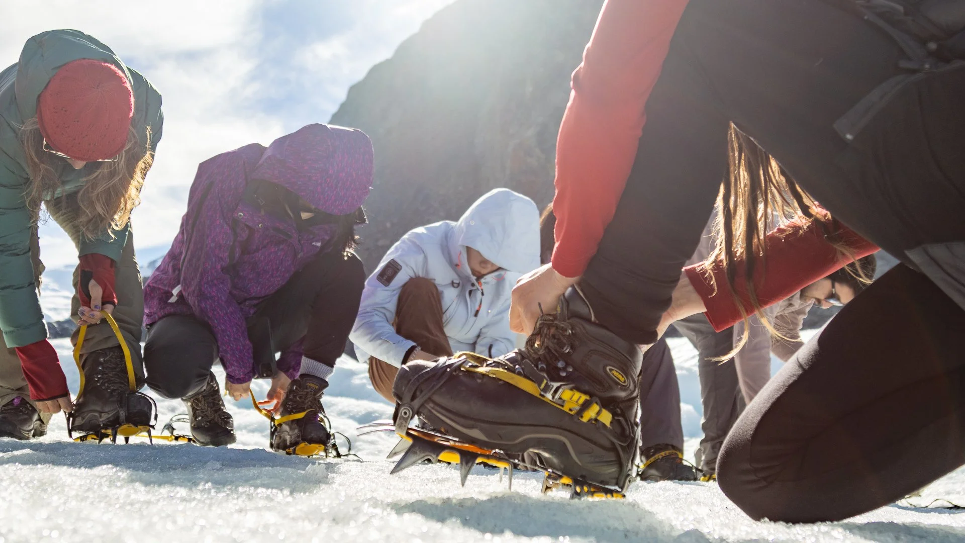 Group of people outdoors in winter, putting on snowshoes on snow-covered ground near mountains.