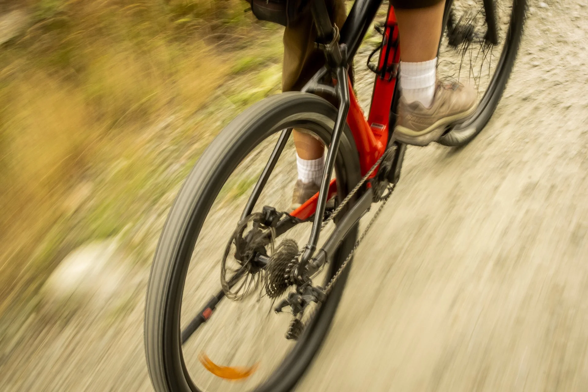 Close-up view of a person riding a mountain bike on a dirt trail, focusing on the bike's front wheel and the rider's legs in beige shoes and white socks.