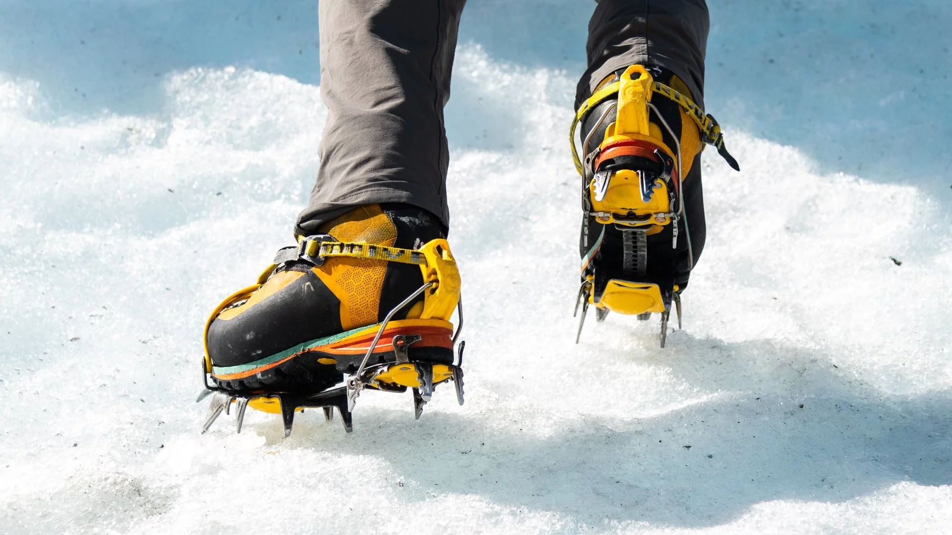 Close-up of a person wearing yellow and black mountaineering boots with crampons walking on snow.