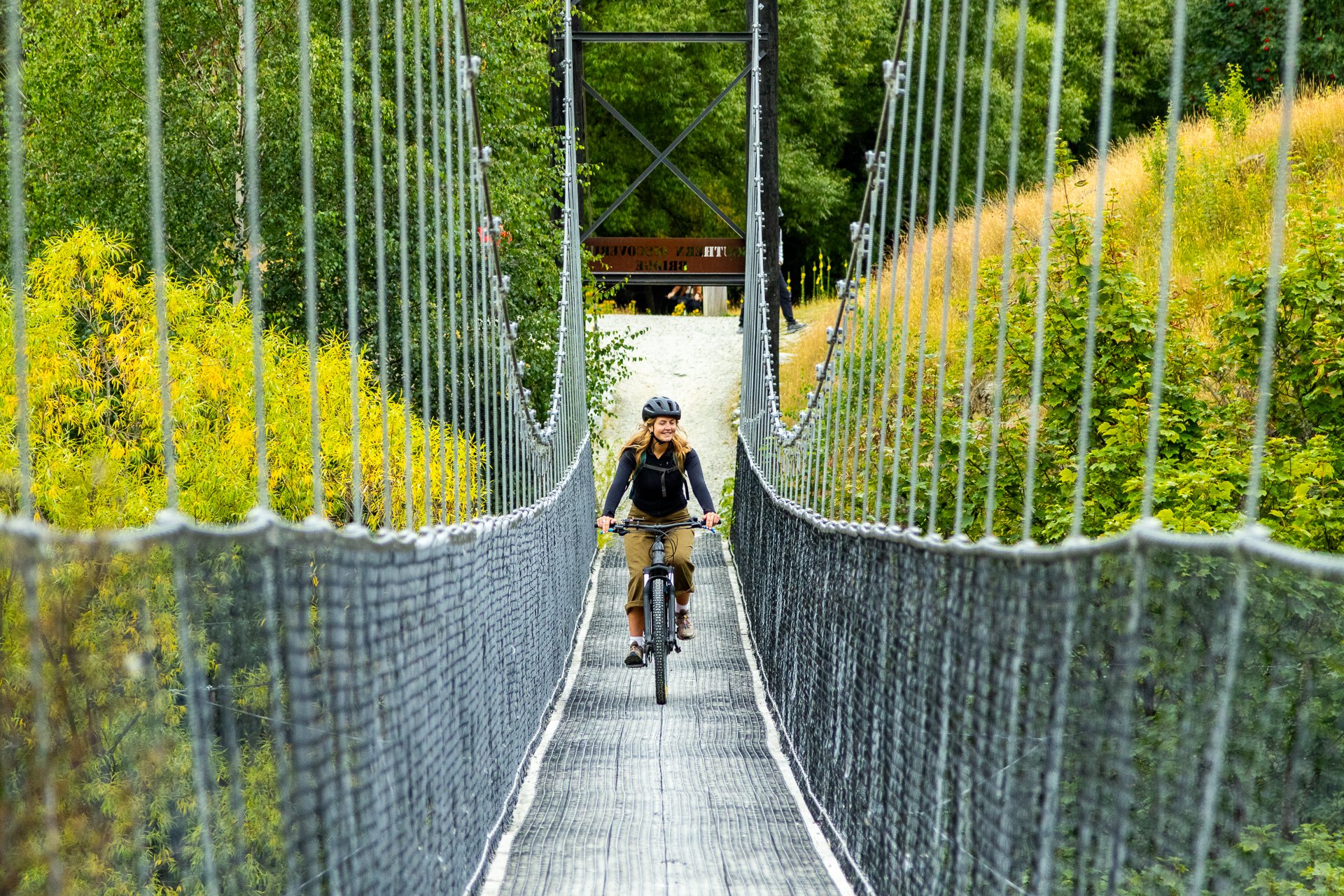 A woman wearing a helmet is riding a bicycle across a suspension bridge with metal mesh flooring and safety netting on sides, surrounded by green trees and plants.