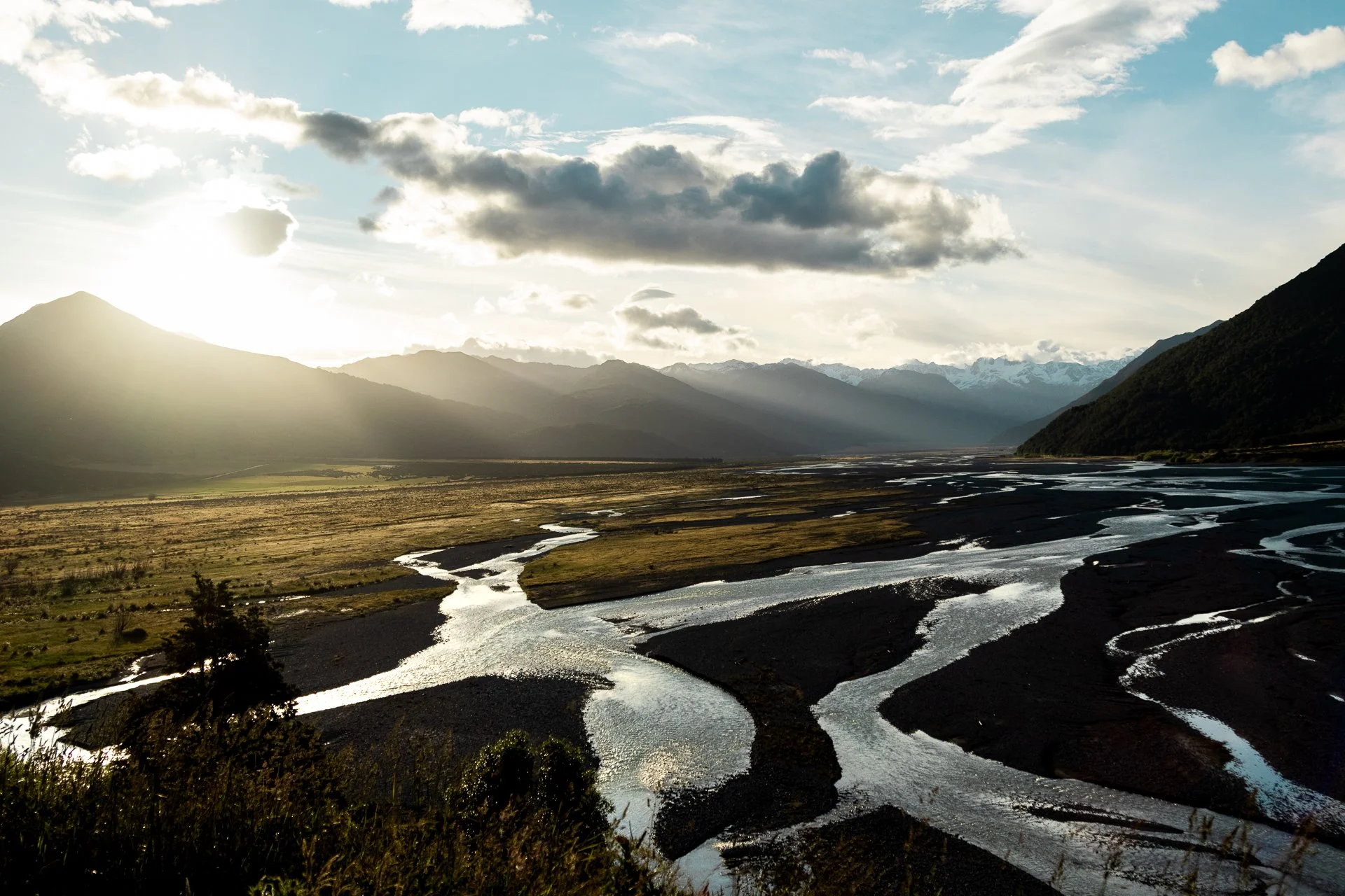 Sunlit mountain landscape with a meandering river flowing through a valley, with mountains in the background and a partly cloudy sky.