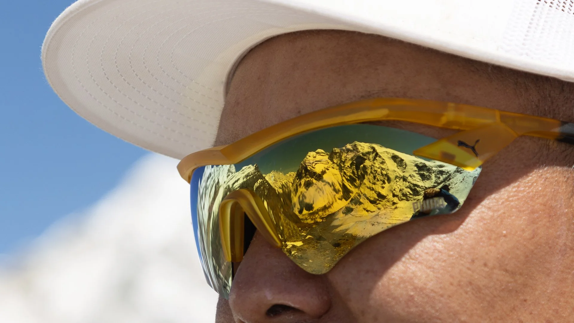 Person wearing yellow reflective sunglasses with mountain reflection, white hat, and tan skin, outdoors with blue sky and clouds.