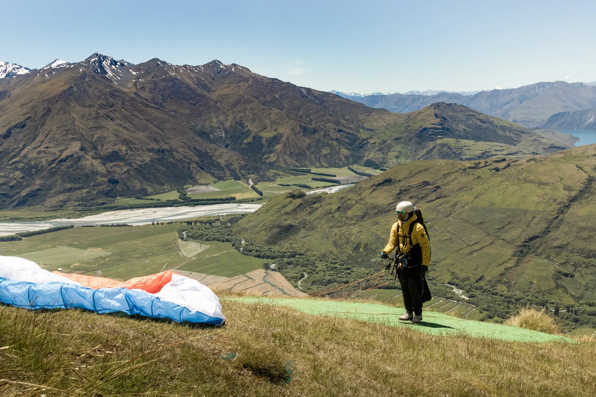 A person prepares for paragliding on a grassy hillside with mountains in the background, wearing a helmet and harness.