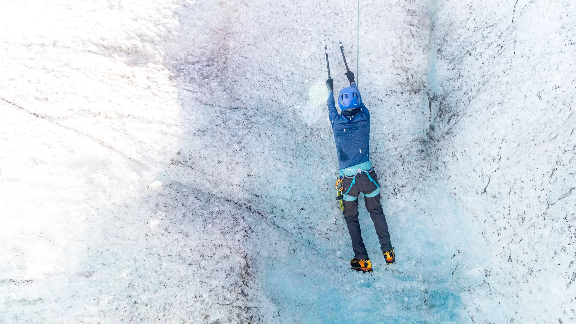 A person wearing a blue helmet and mountaineering gear climbing a snowy ice wall using ice axes.