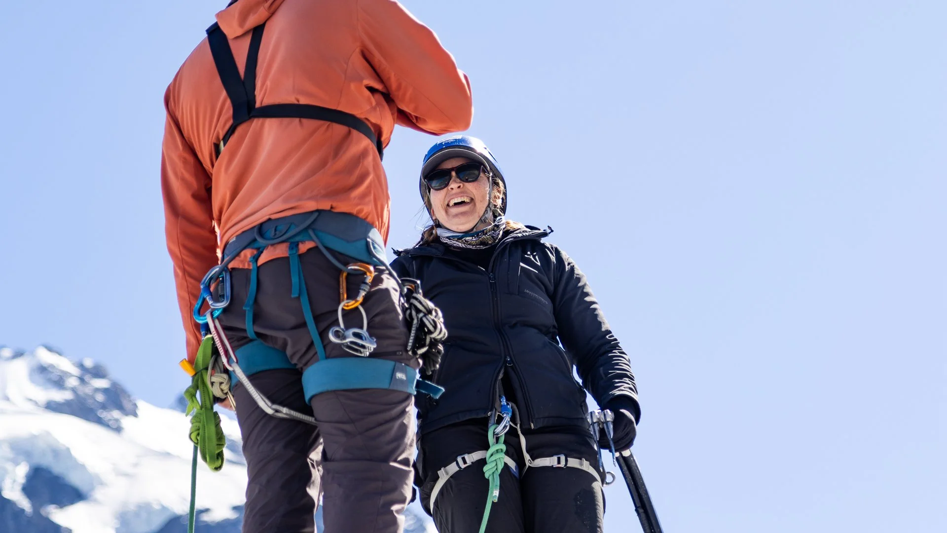 Two climbers in winter gear standing on snow-covered mountain with clear blue sky, engaged in conversation, with one smiling broadly.