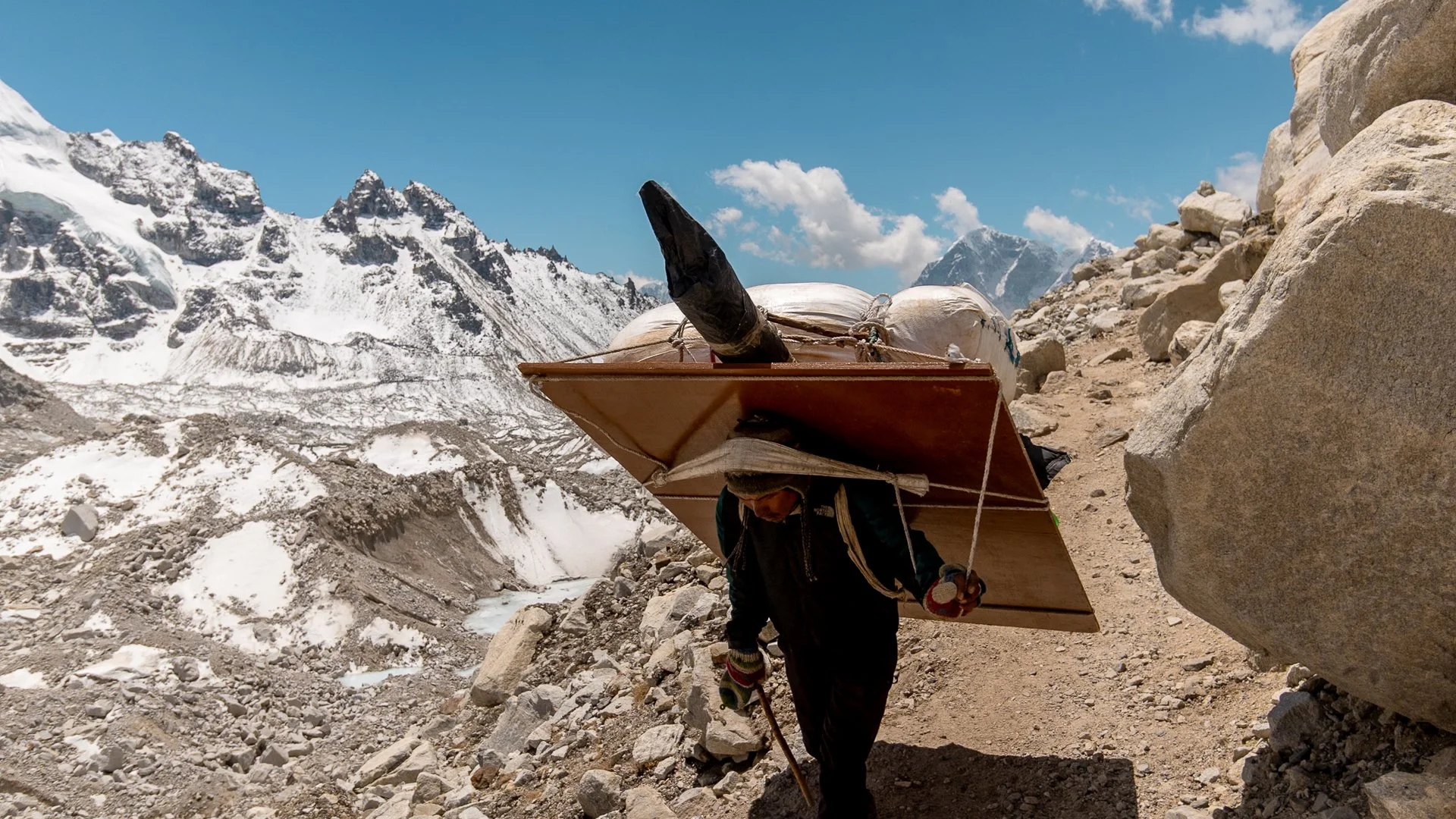 A person hiking in a snowy mountain landscape, carrying a large wooden board and backpack, surrounded by rocks and snow-capped peaks under a partly cloudy blue sky.