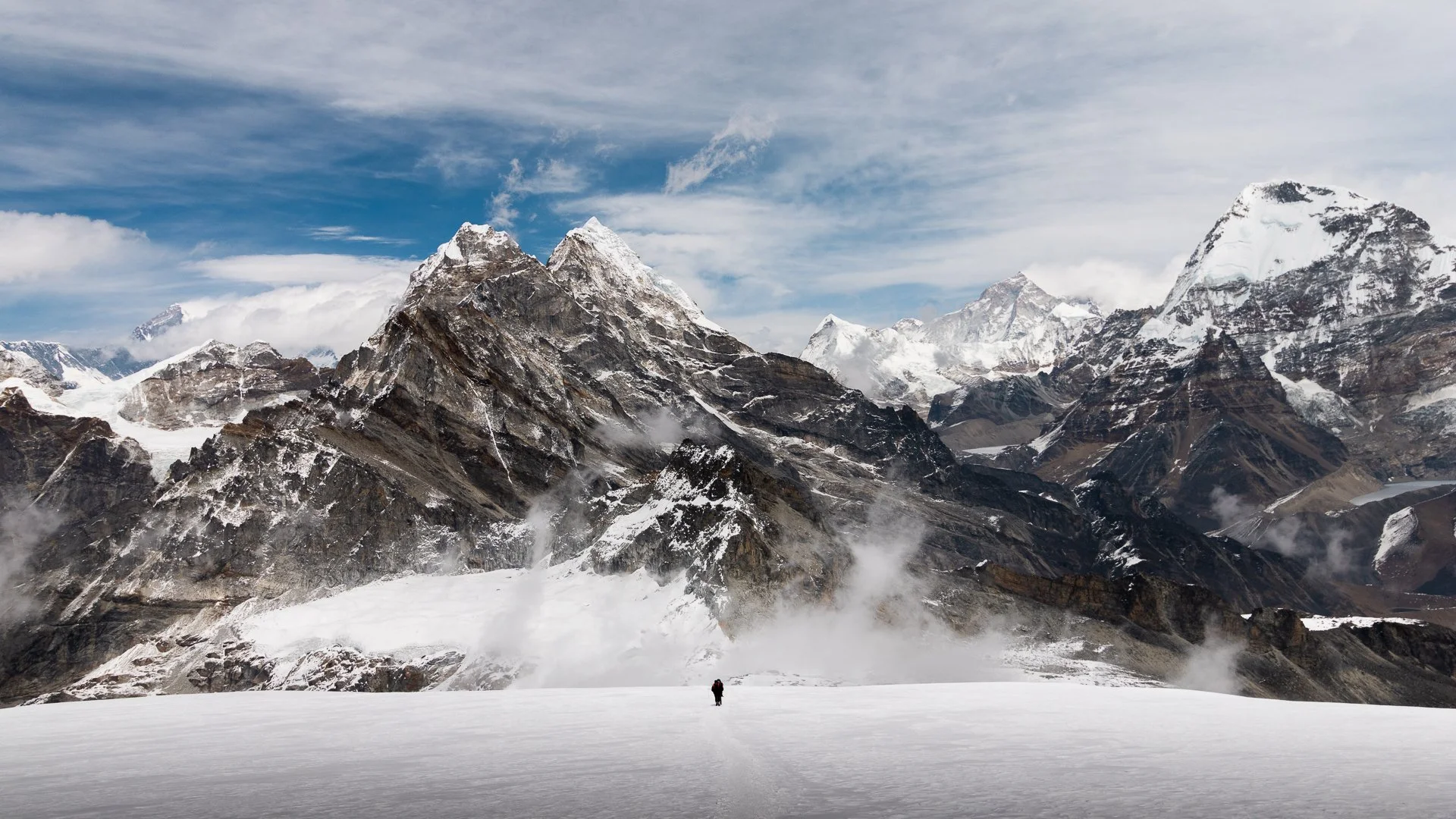 Snow-covered ground with a lone hiker, surrounded by towering rugged mountains with snow and ice, under a partly cloudy sky.