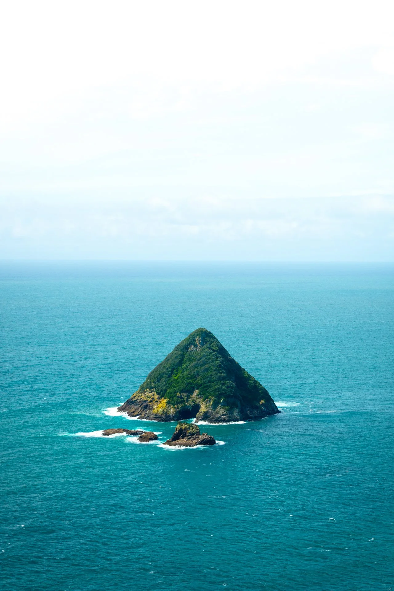 A small, green, rocky island with trees, surrounded by blue ocean, with a cloudy sky in the background.