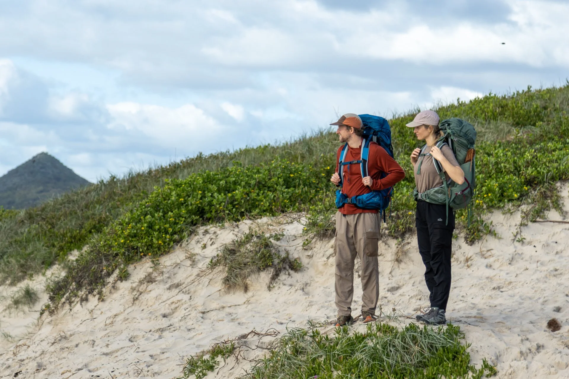 Two hikers standing on a sandy beach with green vegetation, wearing backpacks and hats, looking towards the horizon on a cloudy day.