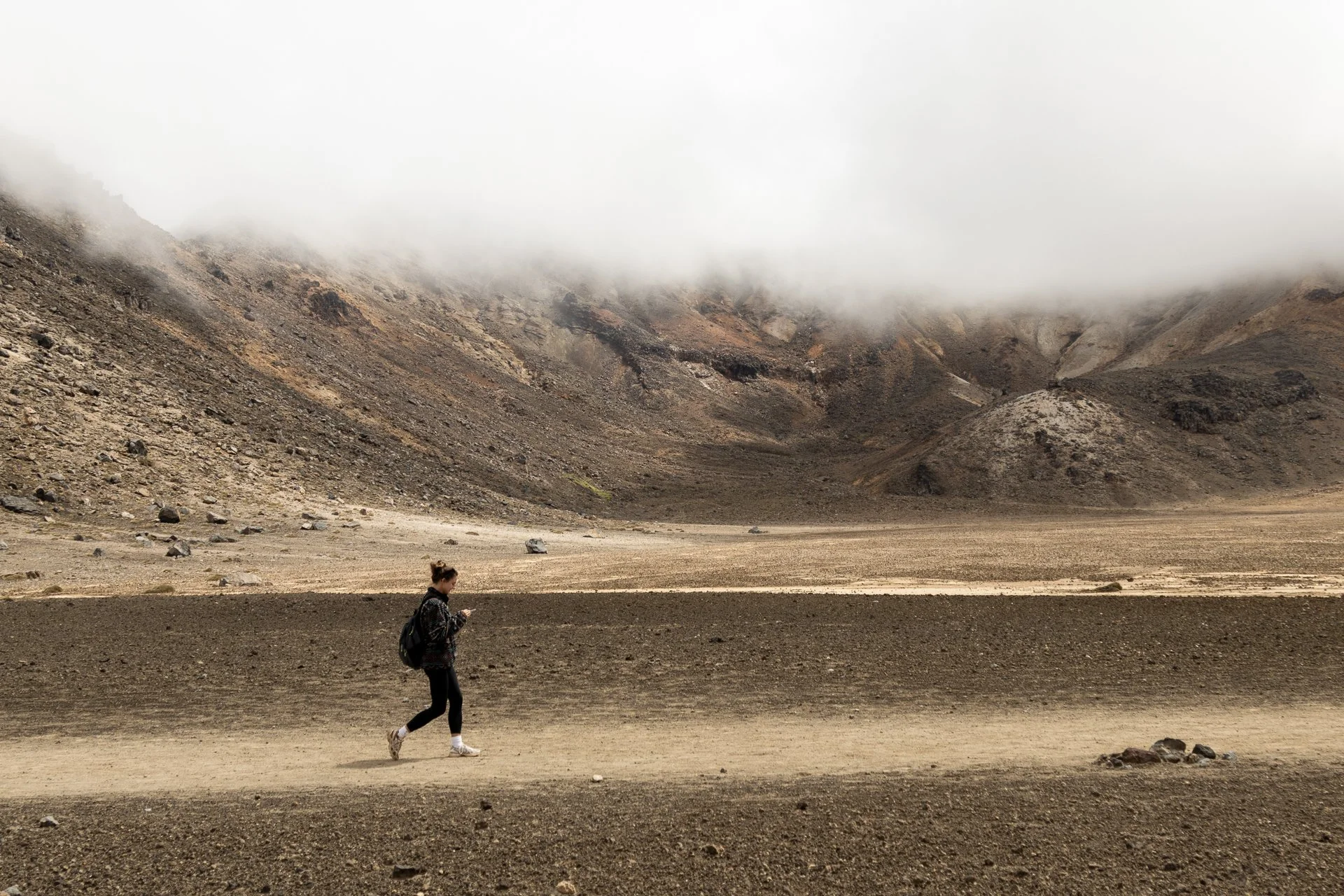 A woman with a backpack walking in a barren volcanic landscape with rocky hills and low clouds in the distance.