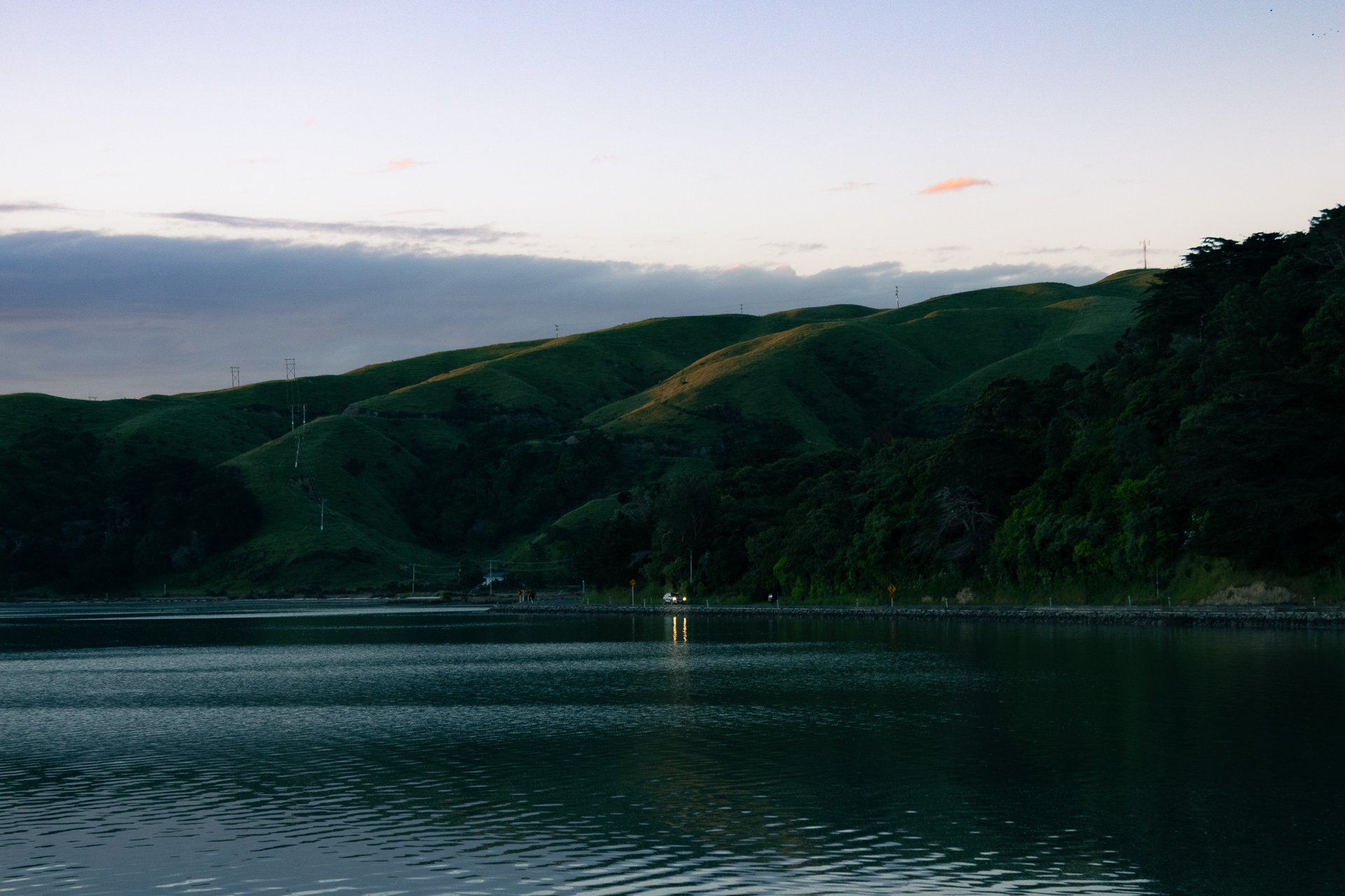 Calm water reflecting green rolling hills under a cloudy sky at dusk.