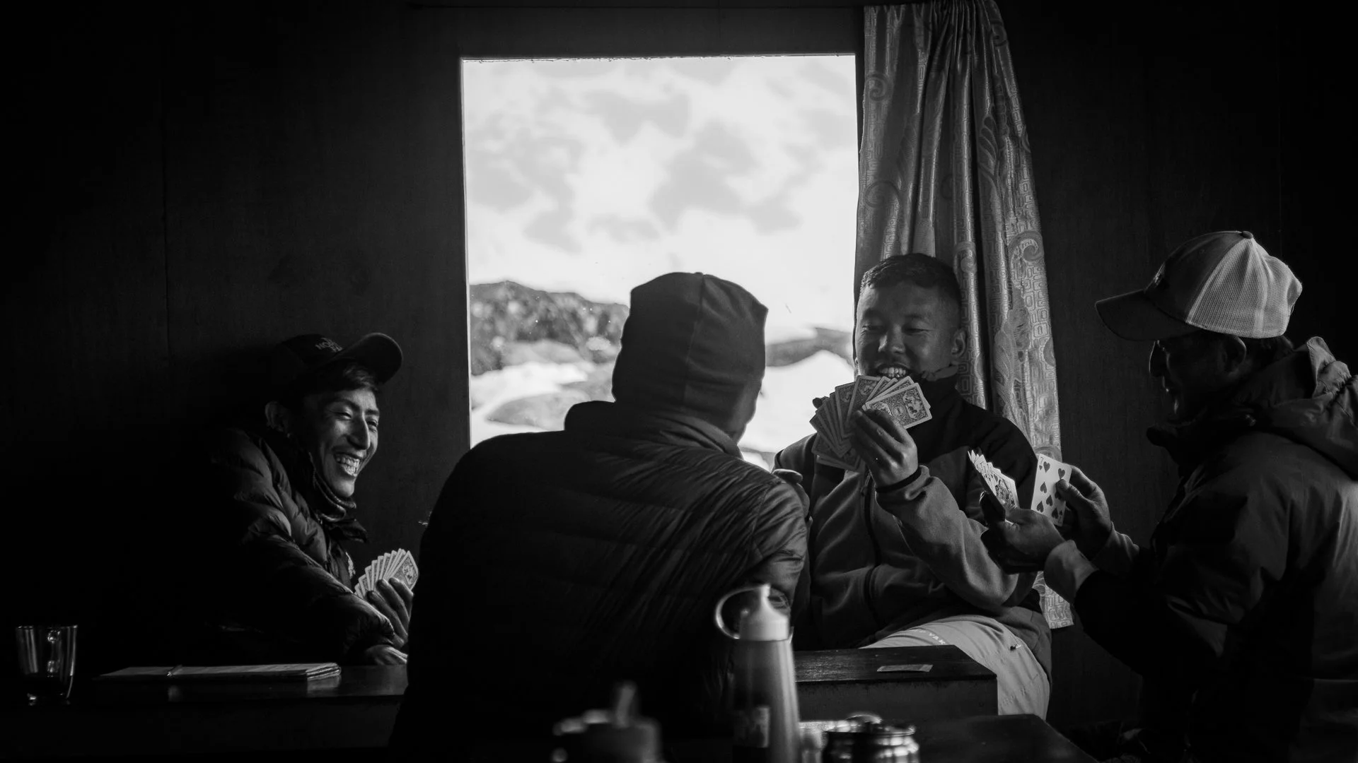 Four men sitting around a table indoors, playing cards and holding money, with one man smiling in the background, in a mountain setting outside the window.