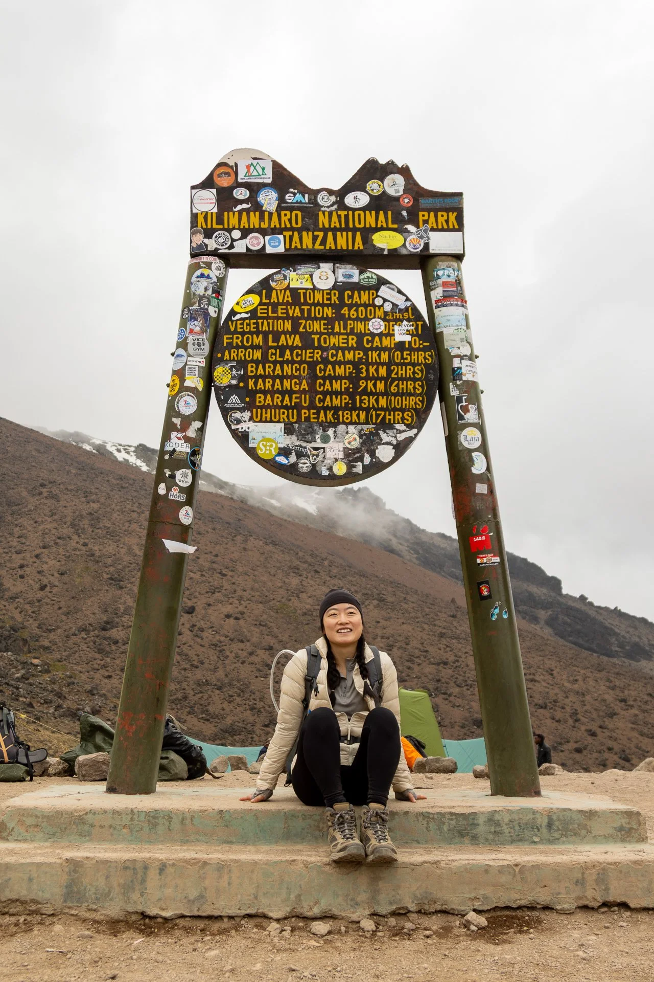 A woman sitting on the ground in front of a large sign at Kilimanjaro National Park, Tanzania, with mountains and volcanic terrain in the background. The sign provides information about the park and nearby campsites.