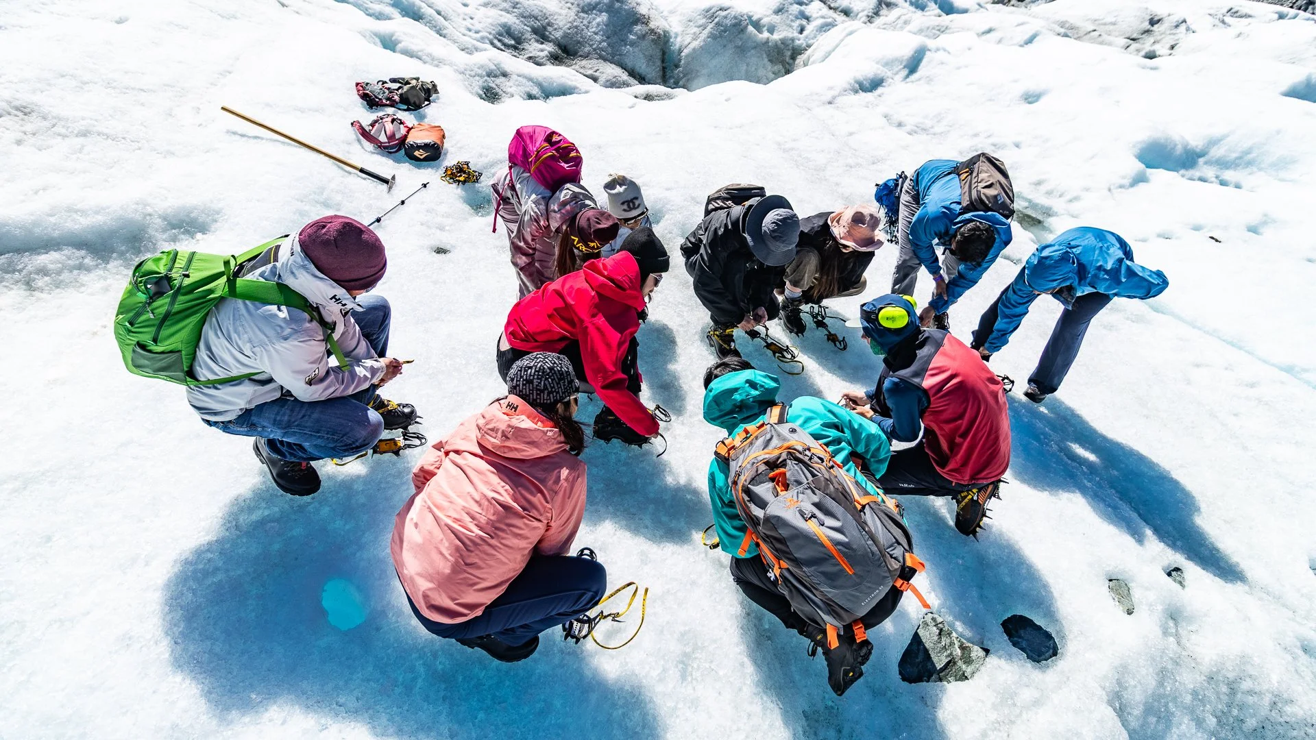 Group of people in winter gear preparing on snow and ice, some kneeling or bending, with backpacks, helmets, and trekking poles.