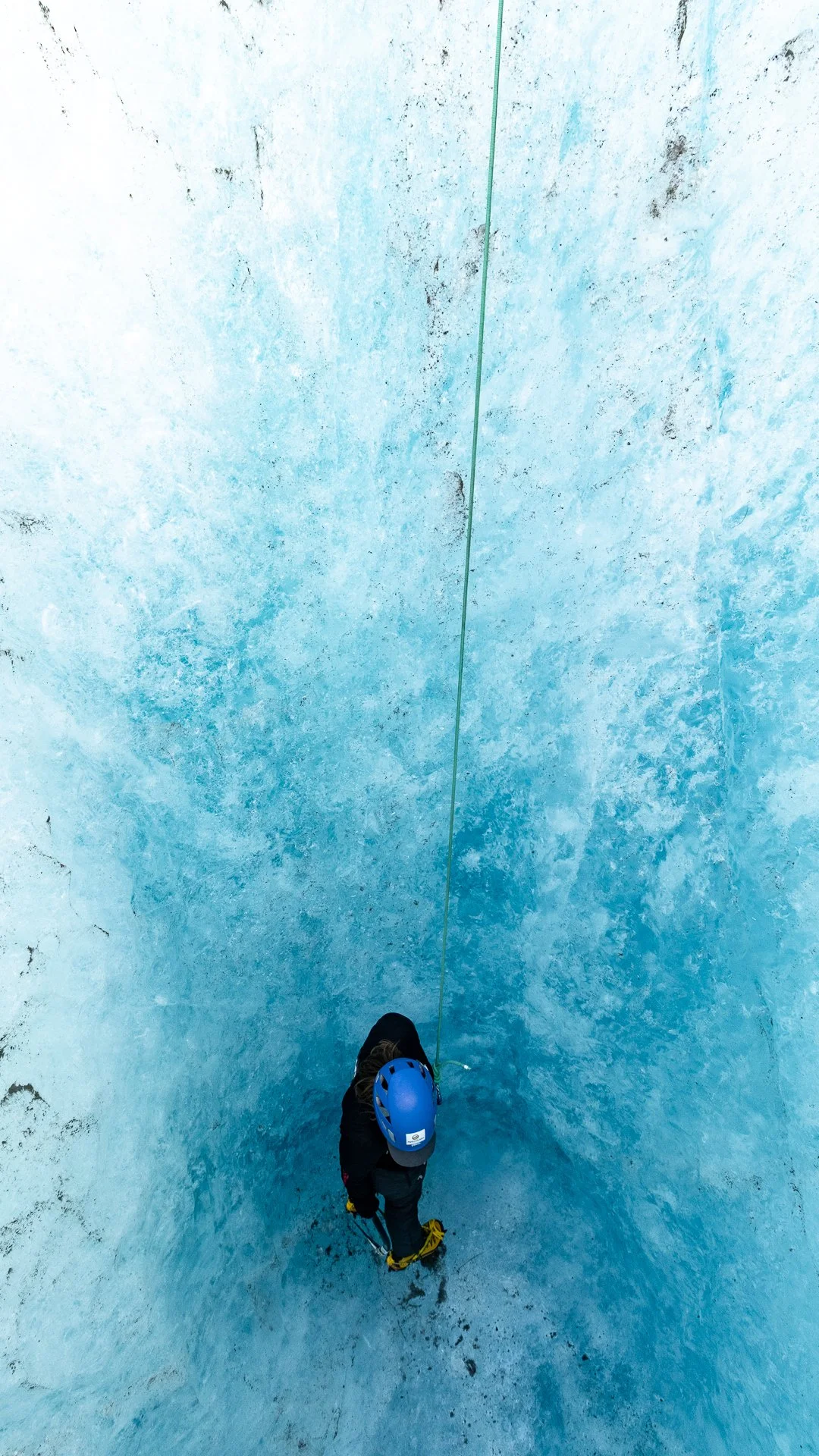 Person wearing a helmet and harness, standing on icy surface, preparing to climb or descend an ice wall, secured with a rope.