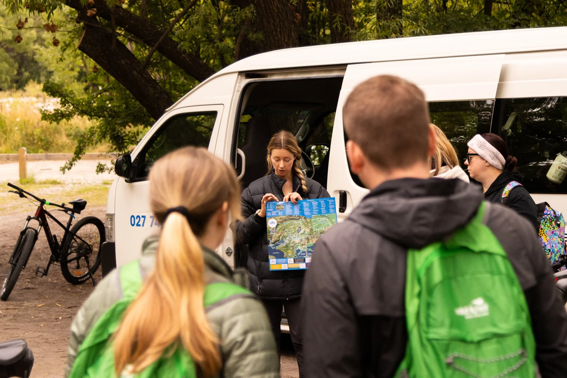 A group of people gathered around a woman who is holding a map and speaking, outdoors near a white van with trees in the background. Bicycles are parked nearby.