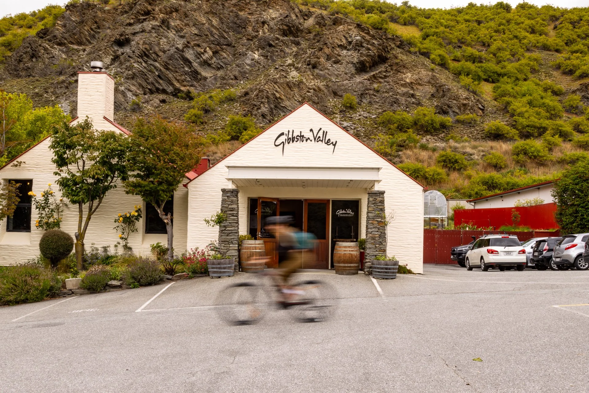 A building with white brick walls and a sign that reads 'Gibston Valley' on the front, located in a parking lot with cars parked and a cyclist passing by. There are green trees and a hillside with sparse vegetation in the background.