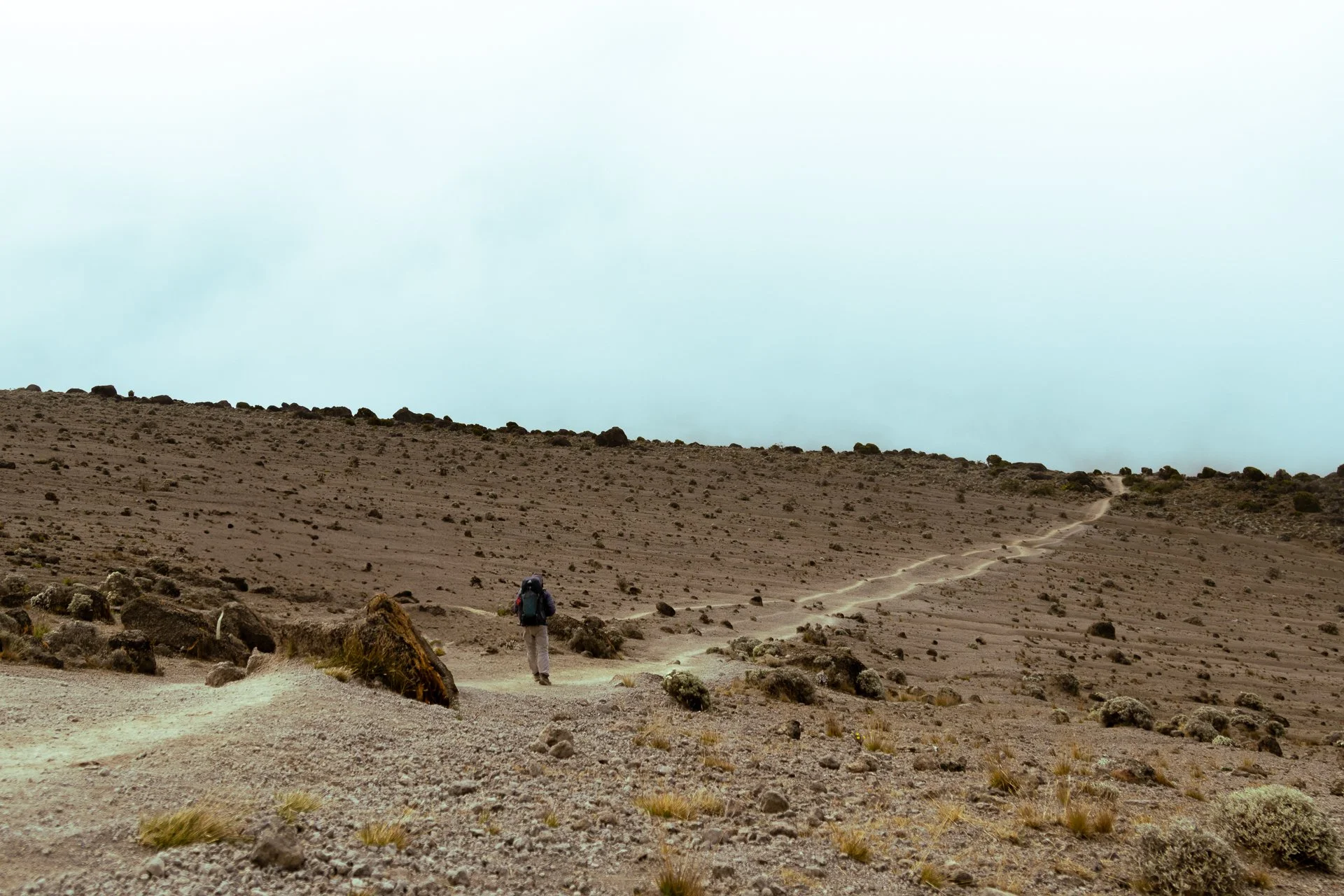 A person with a backpack walking along a sandy trail in a barren, rocky desert landscape under a cloudy sky.