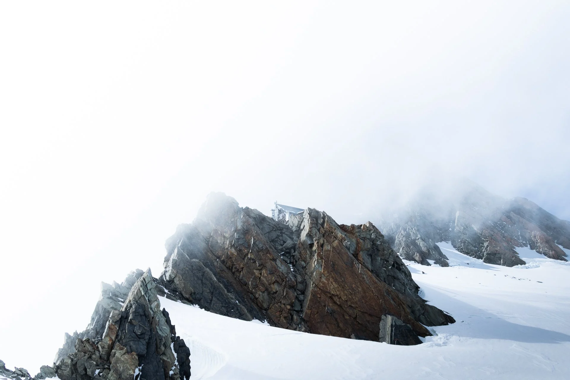 Snow-covered rocky mountain with a distant mountain hut and foggy sky.