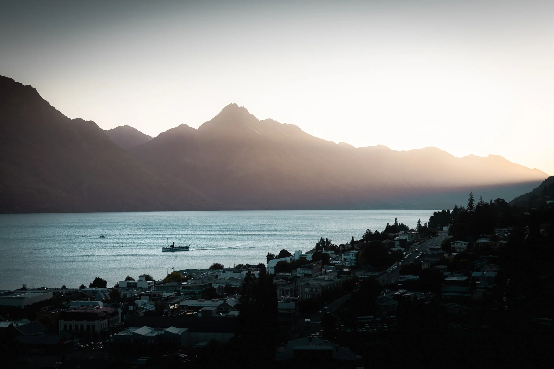 A serene lakeside town with a body of water, mountains in the background, and a boat on the lake, captured during sunset or sunrise.