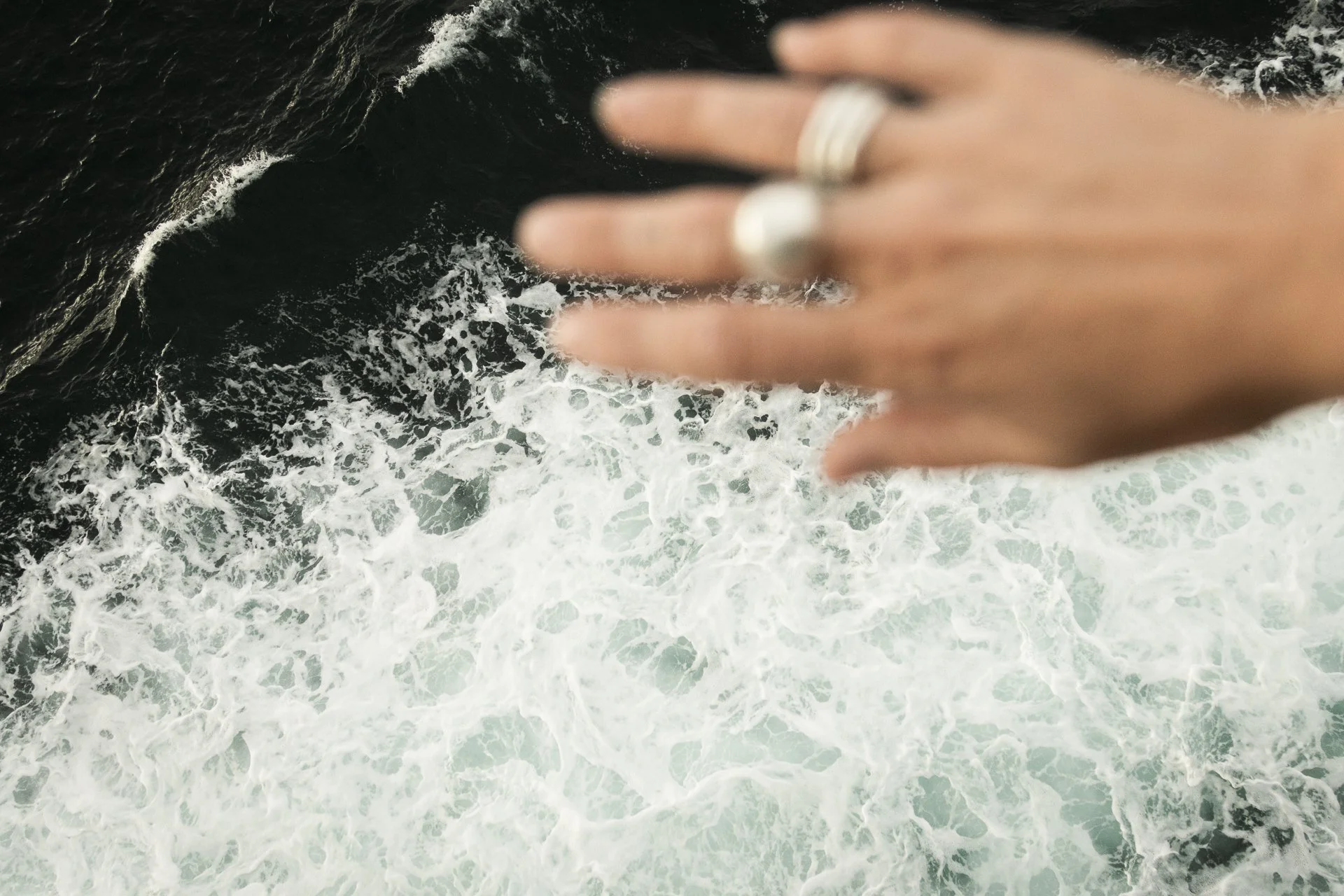 A person's hand with silver rings on the fingers reaching out over the ocean waves from a boat or deck.