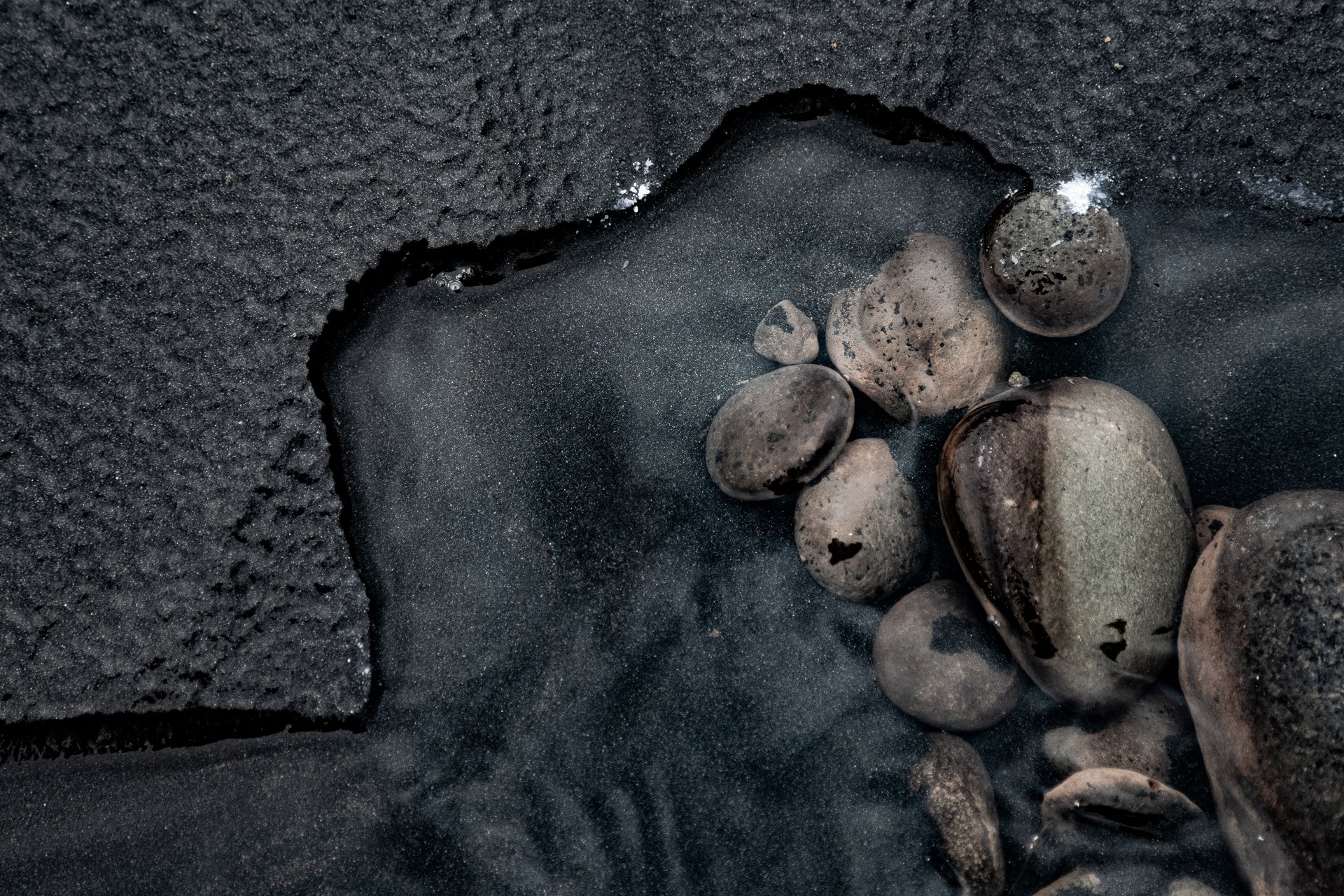 Close-up of dark volcanic sand with a shallow hole containing several small, smooth, rounded rocks and pebbles.