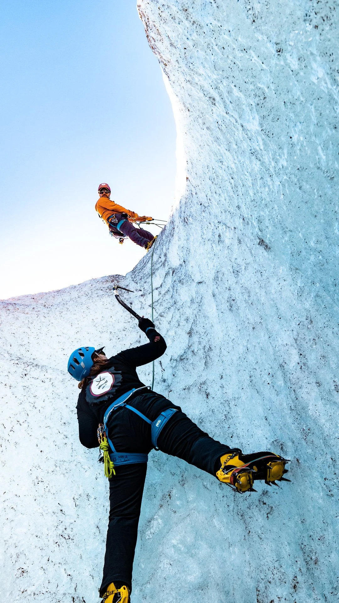 Two ice climbers on an icy cliff, one climbing up and the other assisting from below, both wearing helmets and gear.