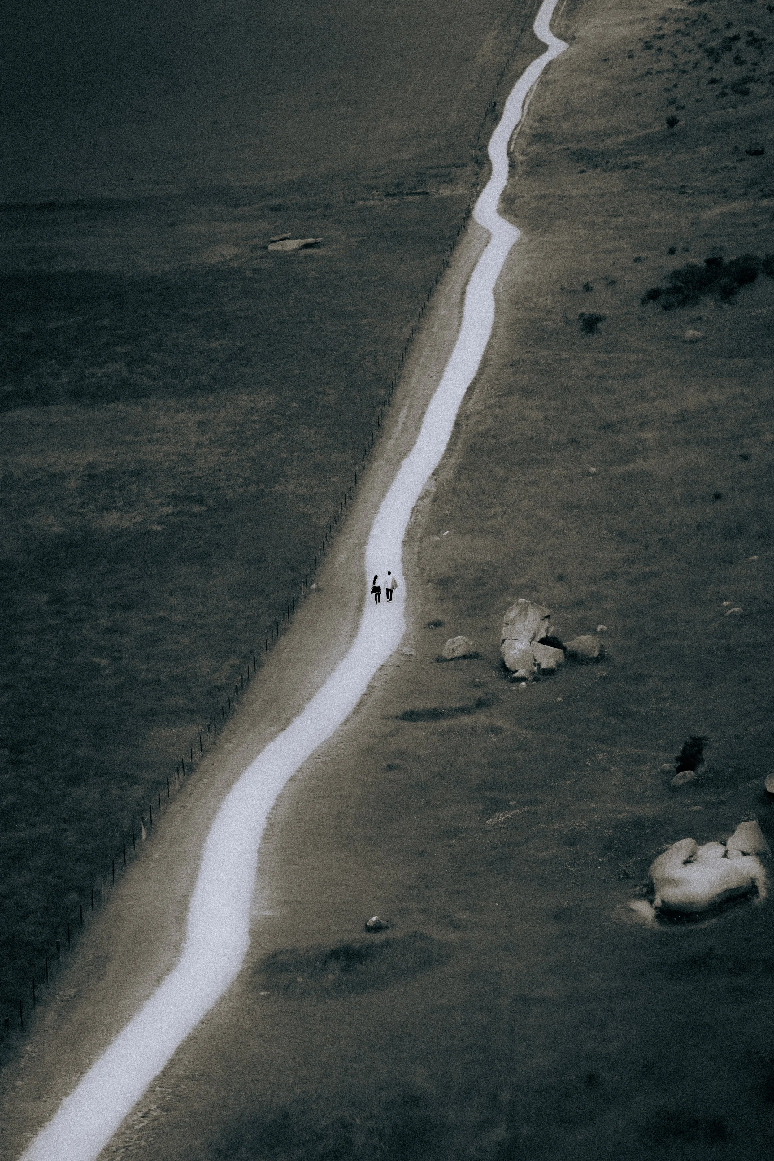 A winding white line runs through a barren, rocky landscape with brown and gray tones. Two small figures are walking along the white line in the distance.