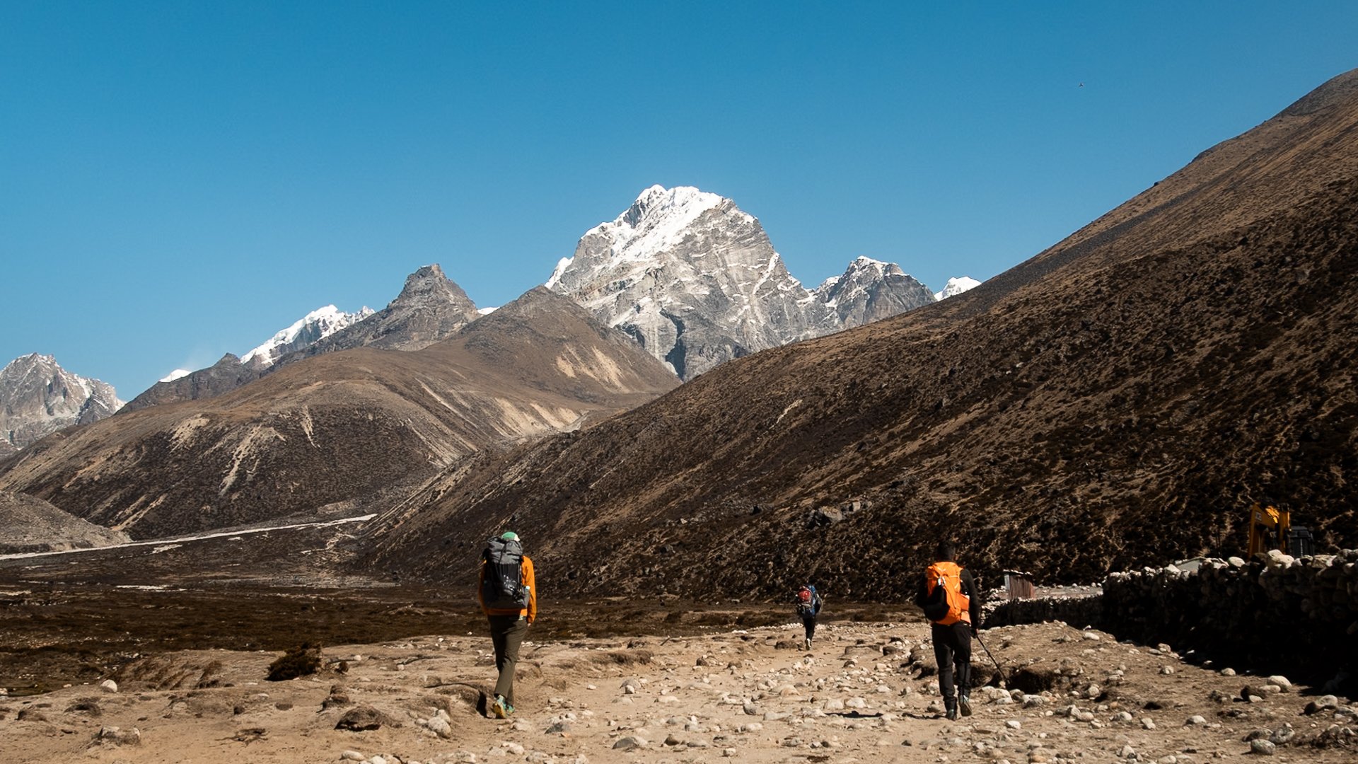 Hikers walking in a mountainous landscape with snow-capped peaks and dry hills under a clear blue sky.