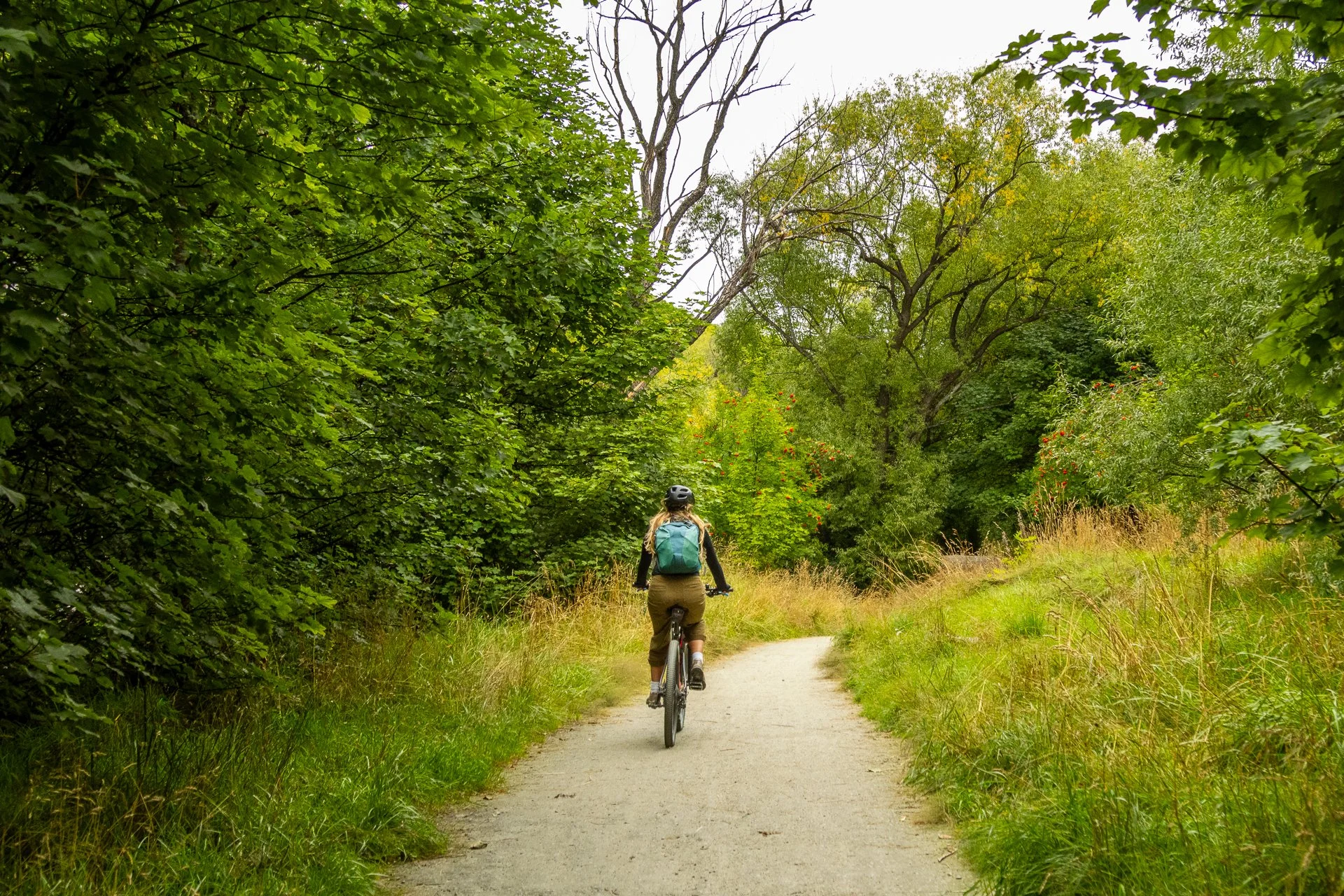 A person riding a bicycle on a dirt path through a lush green wooded area with trees on either side, under a cloudy sky.
