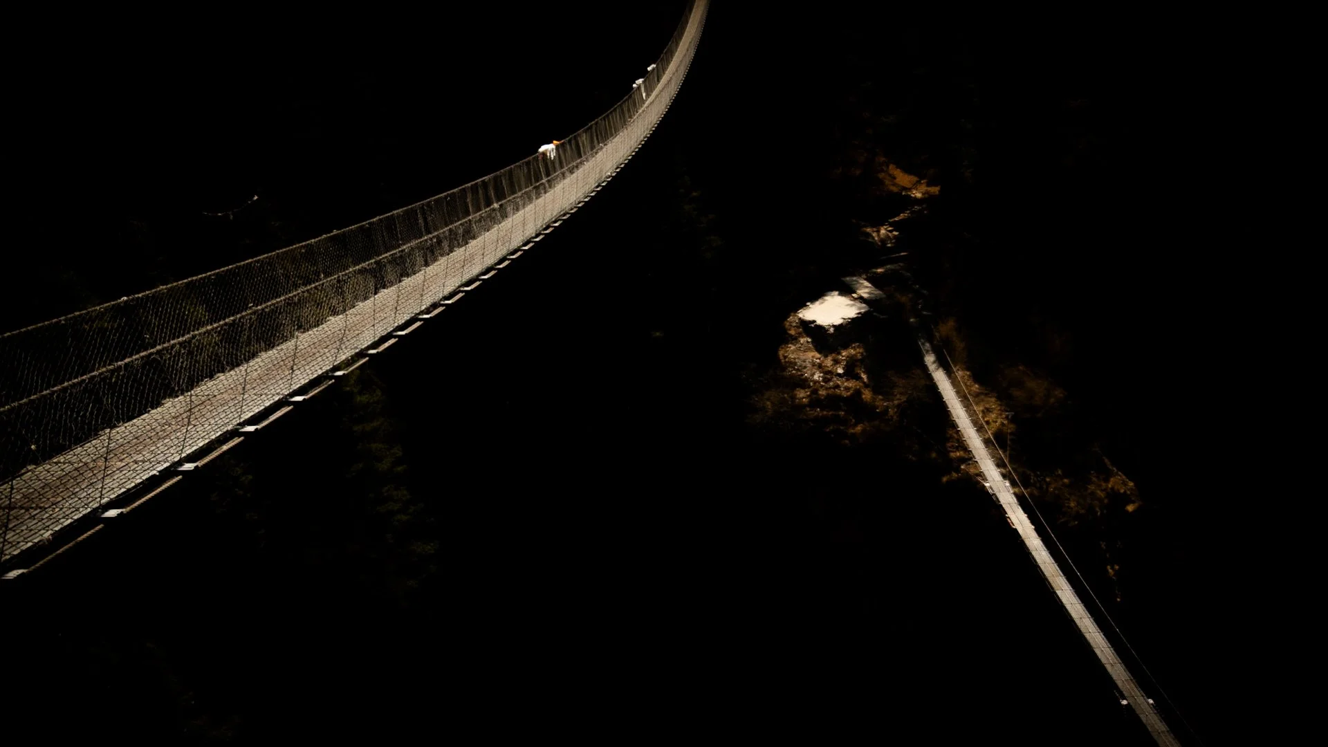 Nighttime view of a long suspension bridge from above, with darkness surrounding and limited lighting along the bridge.