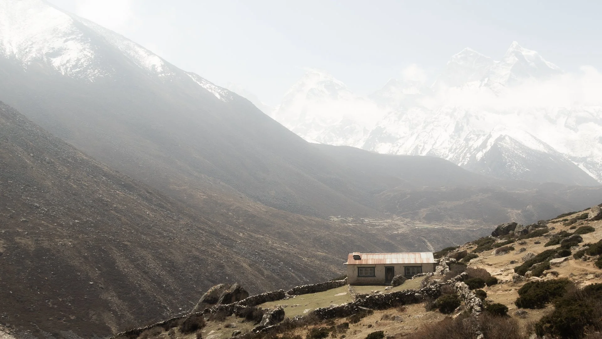 A house with a rusted metal roof in a mountainous landscape with snow-capped peaks in the background and dry rocky terrain in the foreground.