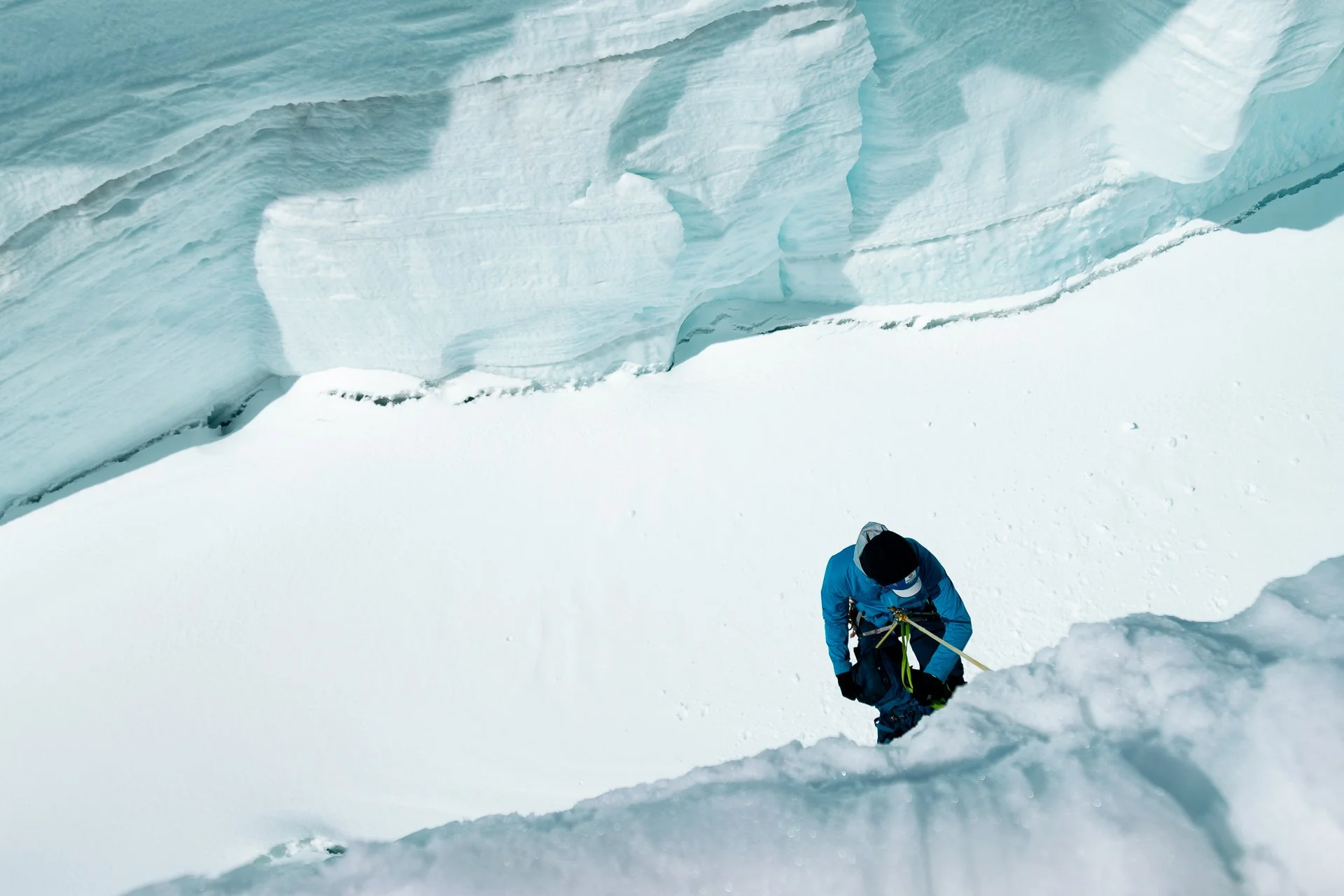 A person in a blue jacket and black hat is climbing a steep, snowy slope, using an ice axe. The scene features large, blue ice formations or glaciers in the background.