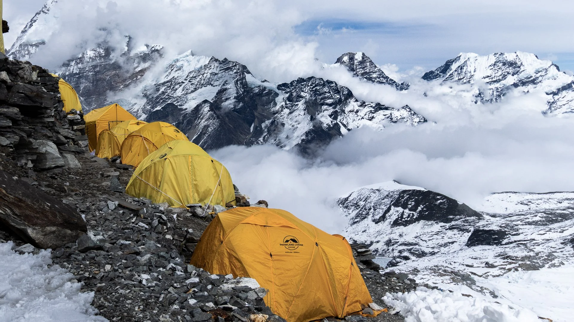 A mountain campsite with yellow tents set up on rocky ground next to a snow-covered slope, with snow-capped peaks and clouds in the background.