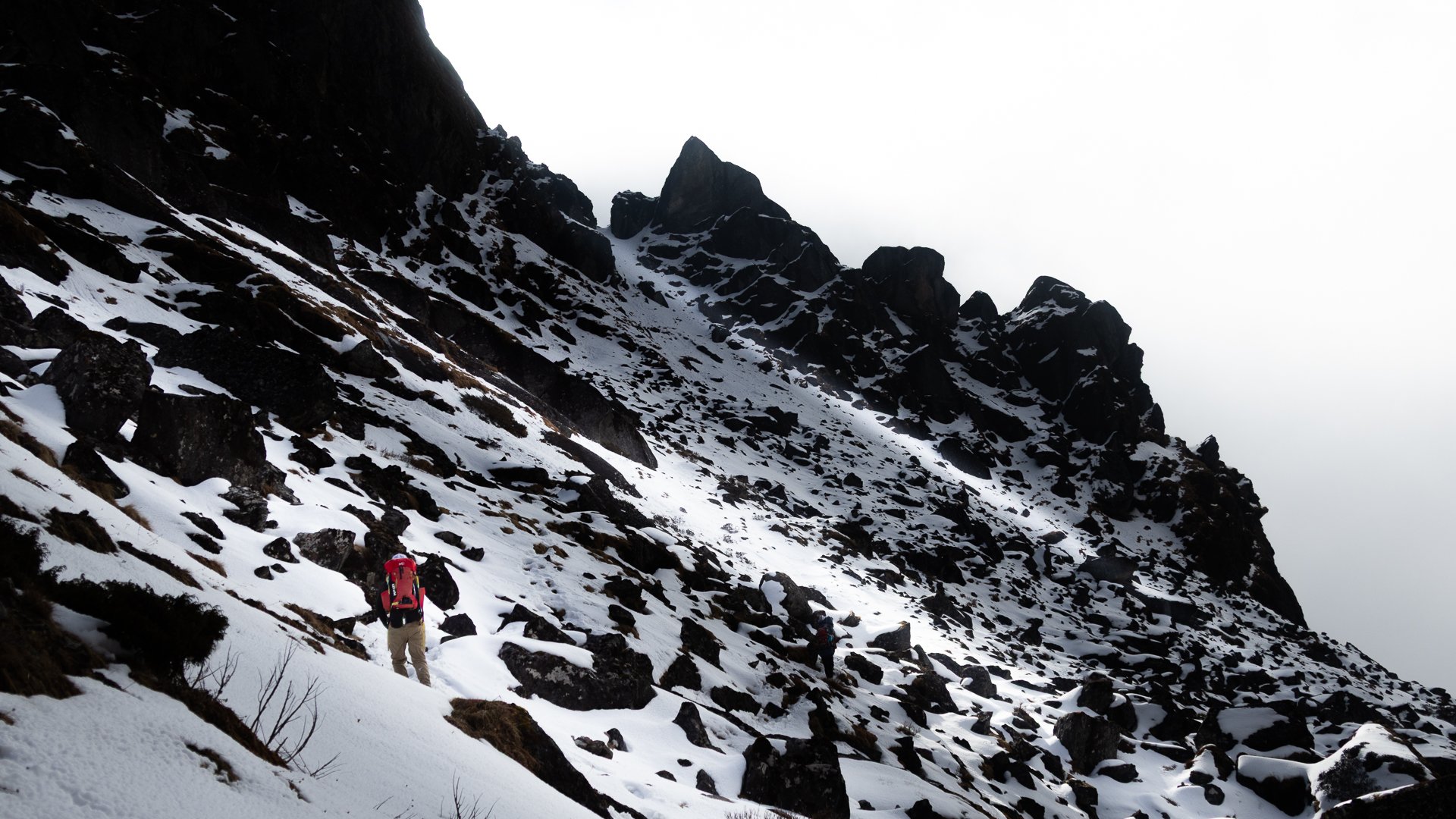 Mountaineers hiking on a snow-covered mountain slope with large dark rocks and a steep, jagged mountain peak in the background.