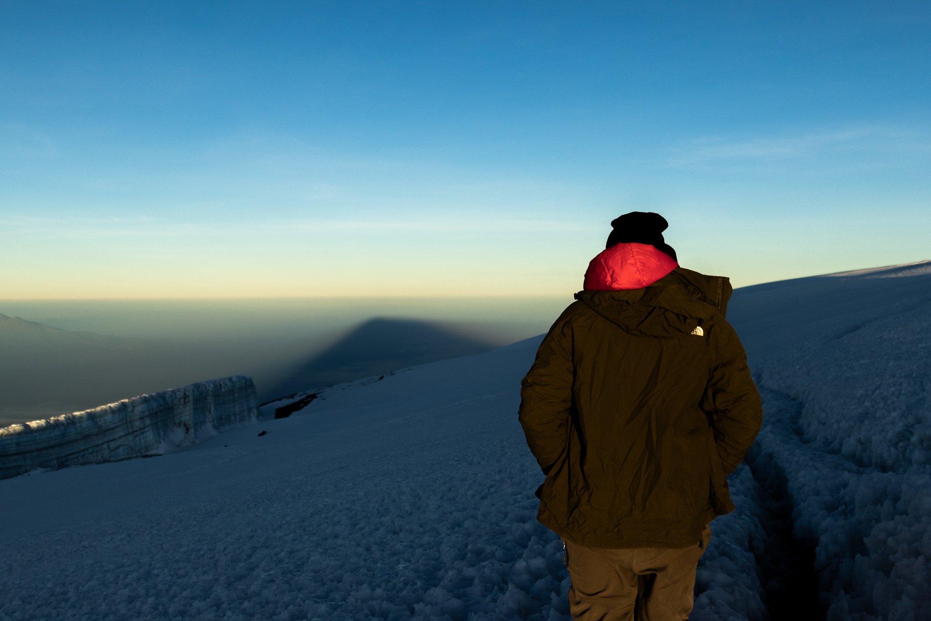 Person wearing a brown jacket and black hat standing on snow-covered mountain at sunset, overlooking a vast snowfield and distant mountains.