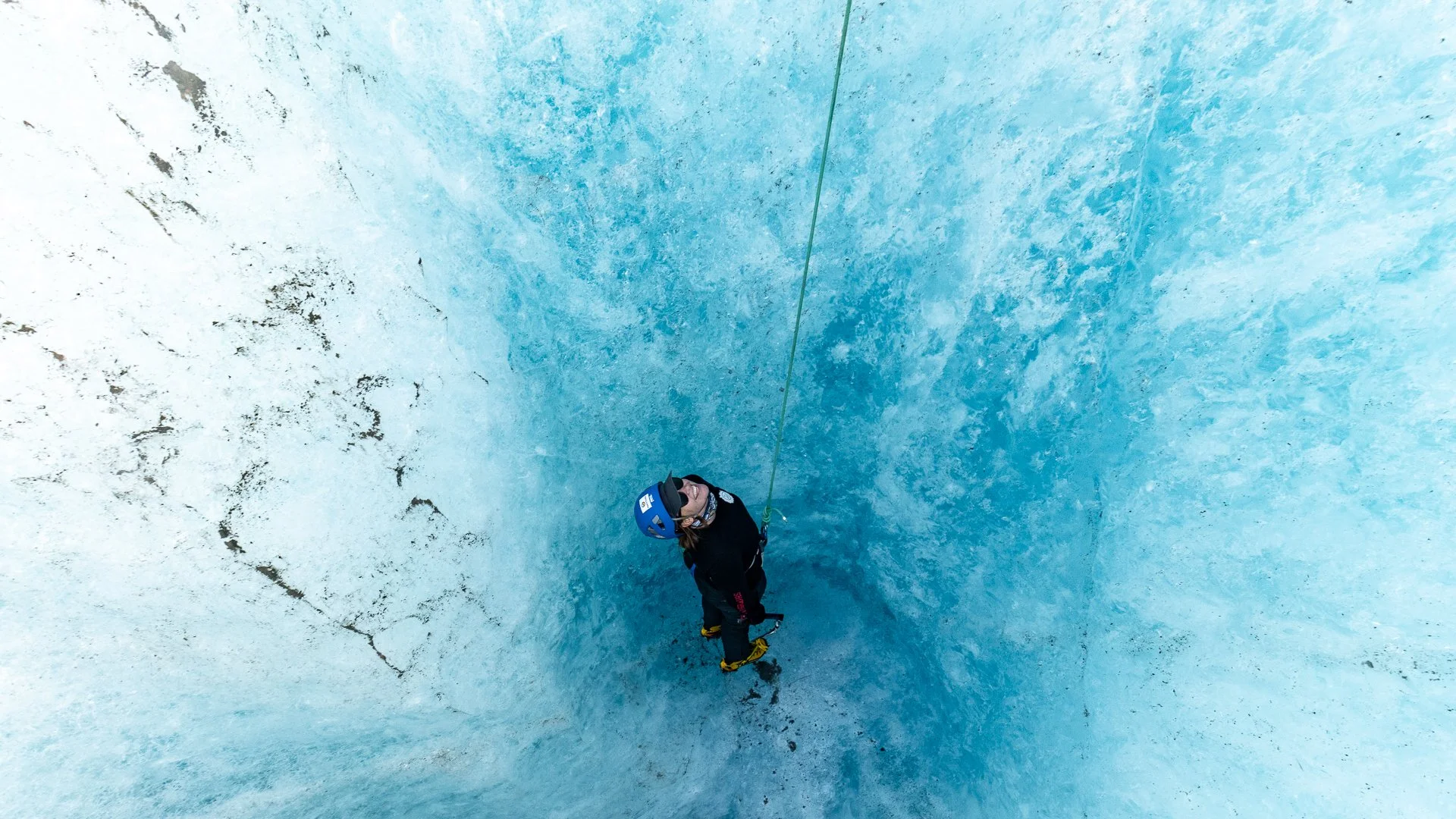Person in a blue helmet climbing or rappelling inside an ice cave or glacier, with wearing ice climbing gear and holding a rope, surrounded by blue ice walls.