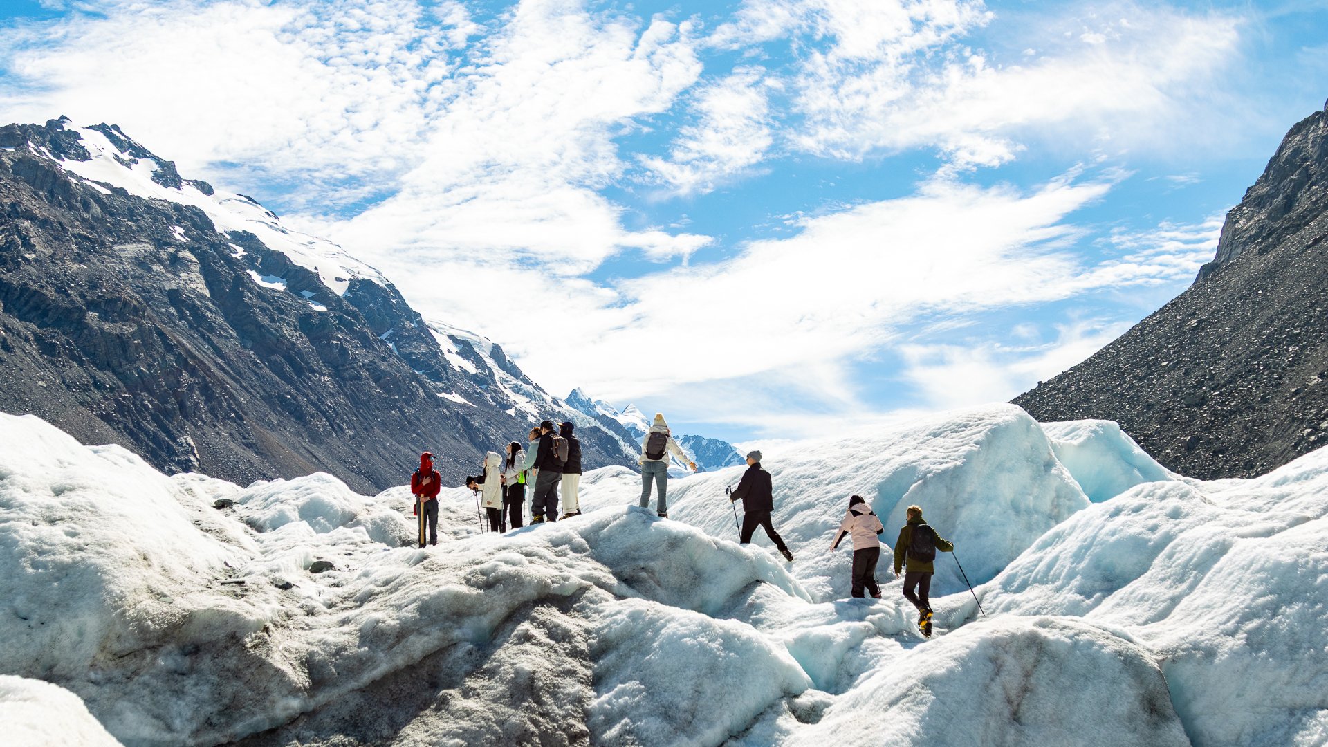 Group of people hiking on snow and ice in a mountainous, scenic landscape with snow-capped peaks and partly cloudy sky.
