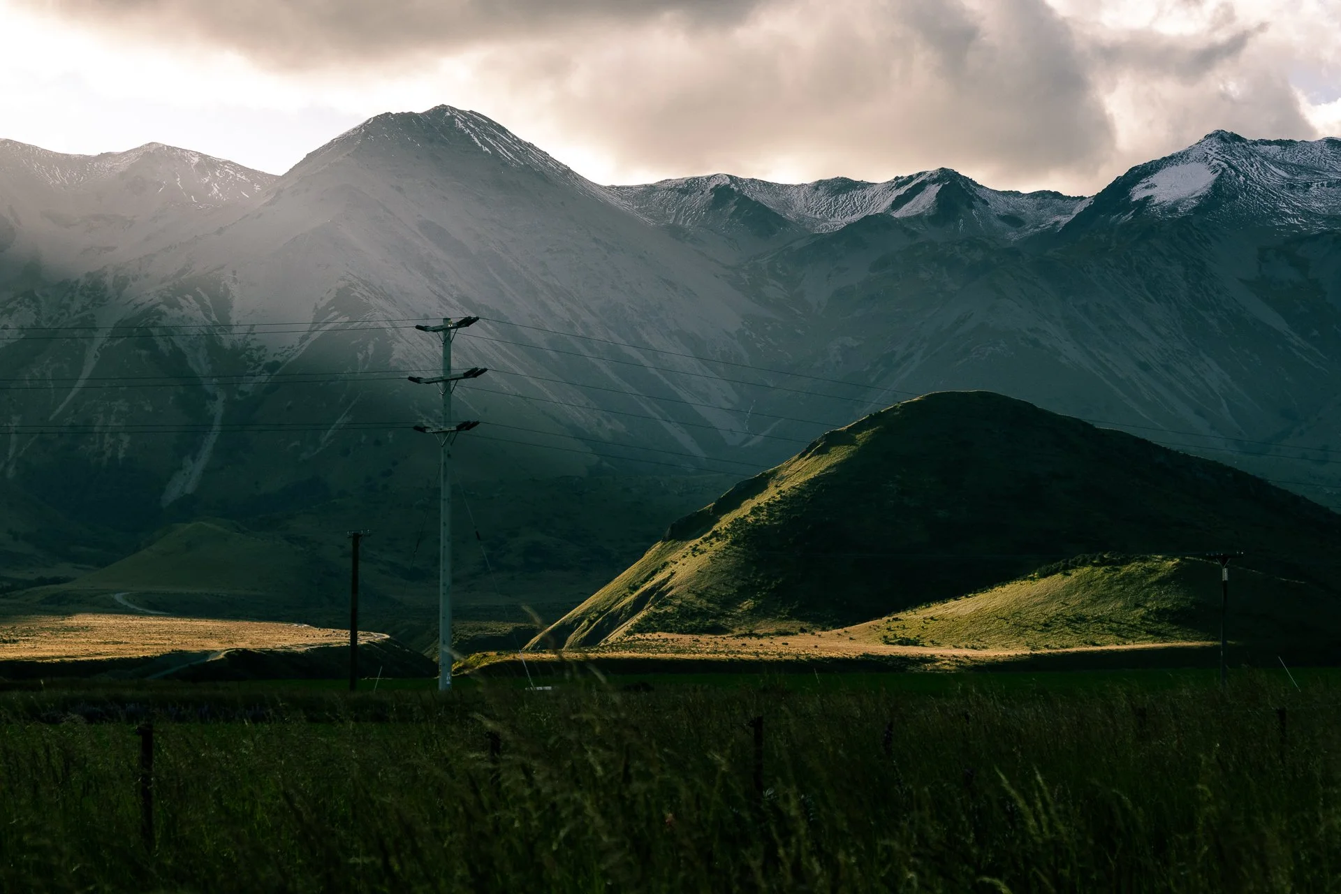 Mountains with snow patches, rolling green hills, and electrical poles in the foreground under a cloudy sky.