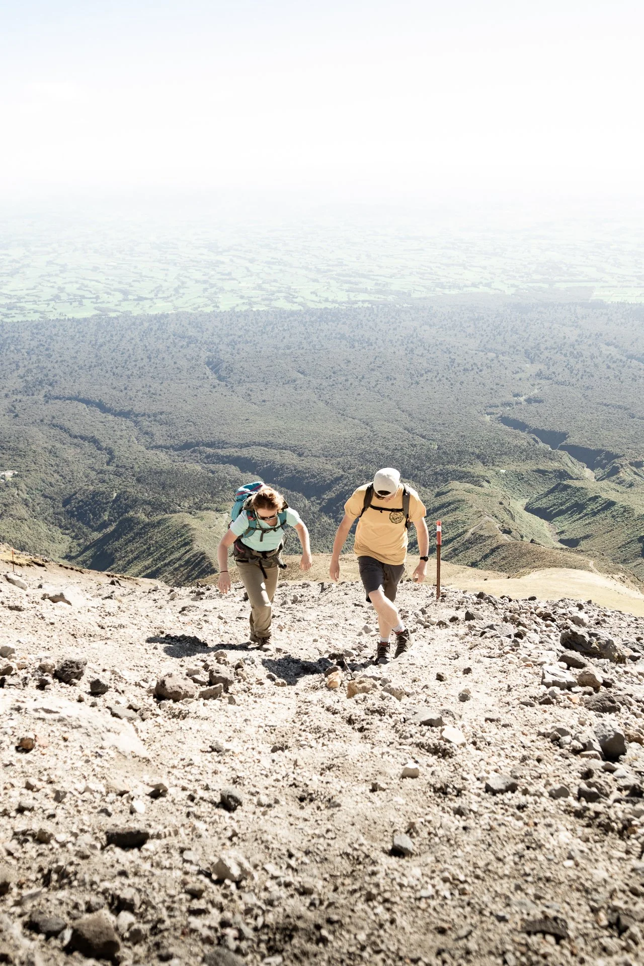 Two hikers ascending a rocky mountain trail with a scenic valley below and distant hills.